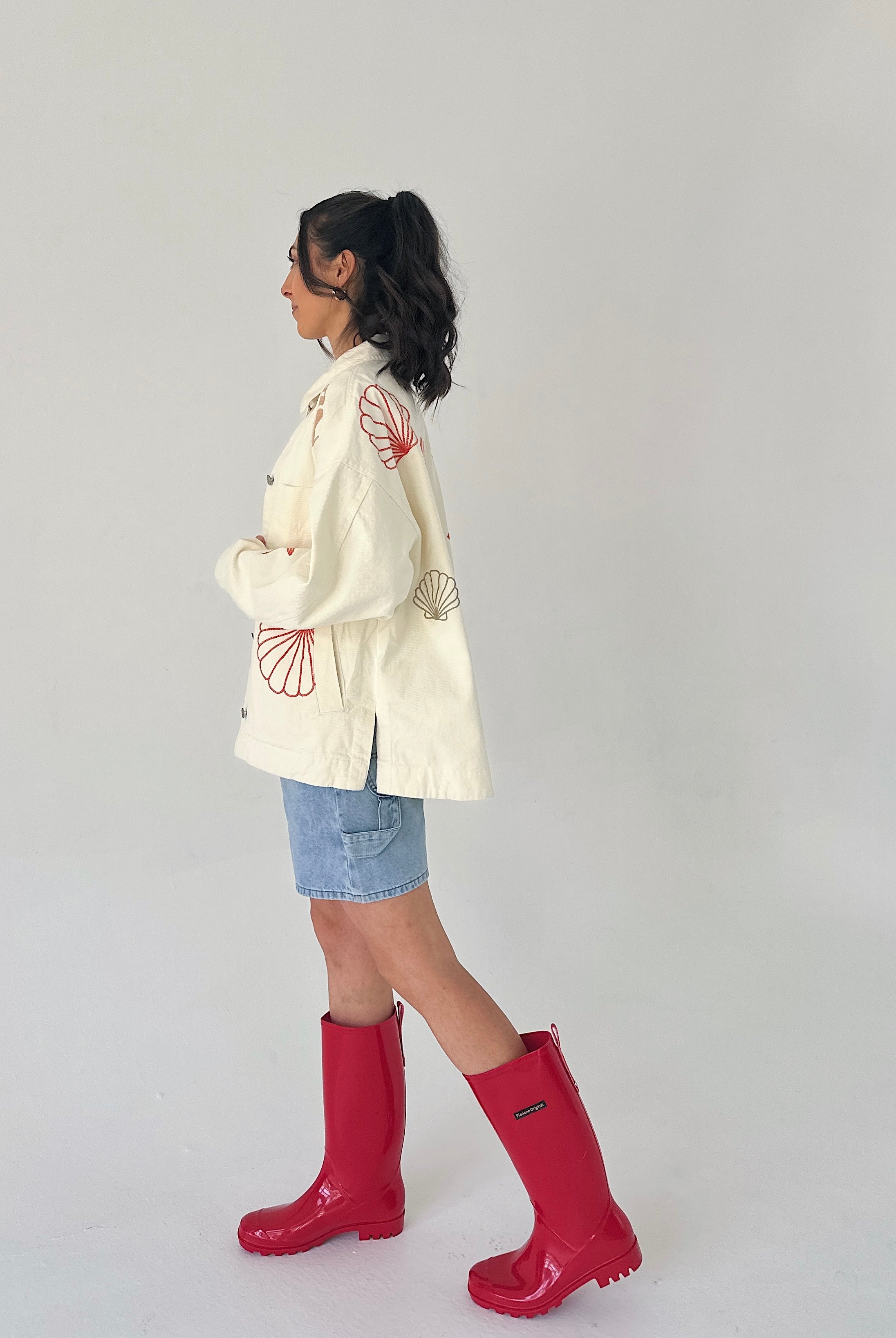Person wearing a white jacket with red patterns, denim shorts, and red rain boots on a plain background