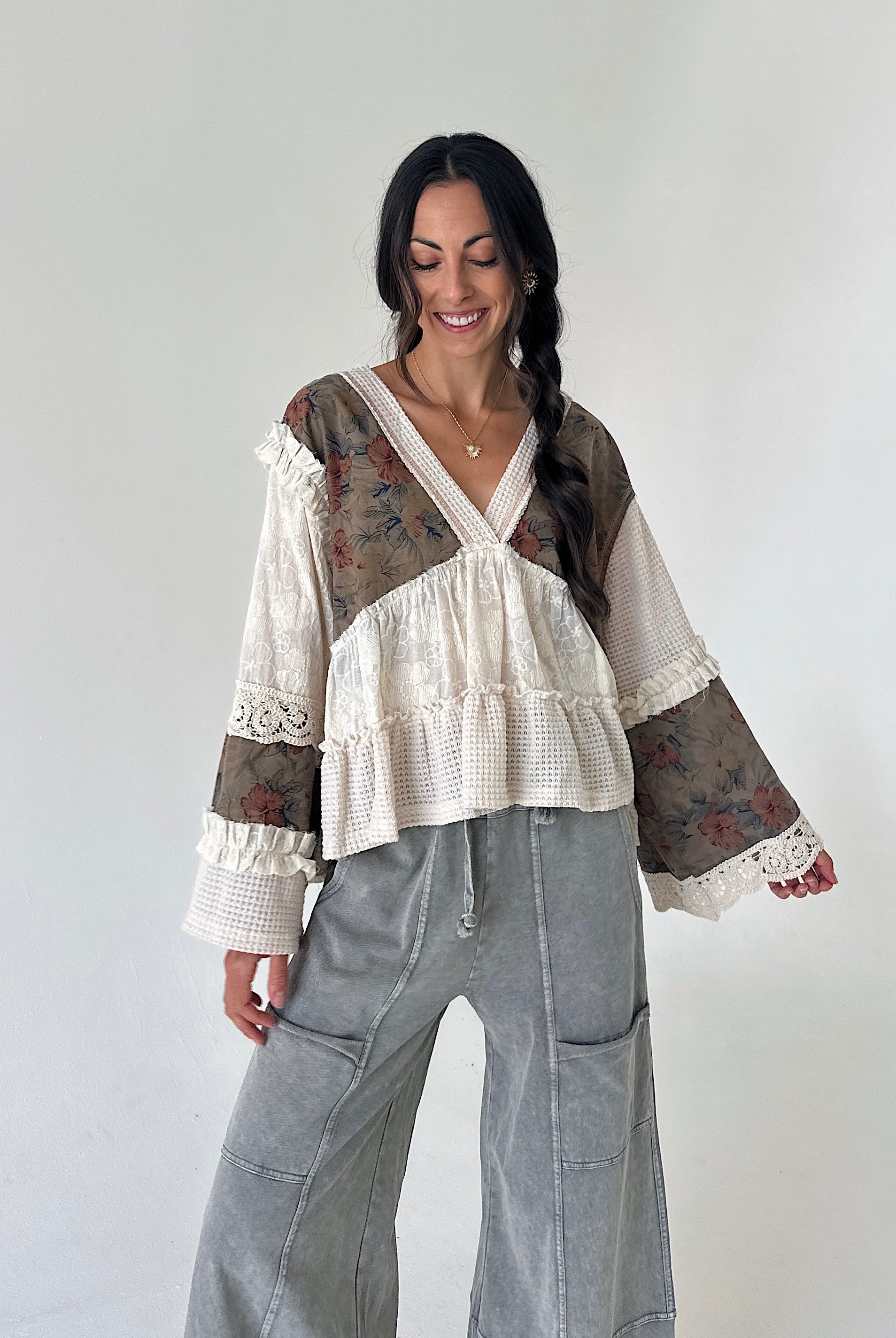 A woman modeling a bohemian-style long-sleeve blouse with cream waffle-knit fabric, lace trim, and brown floral patterned panels, paired with wide-leg gray pants, against a plain off-white background.