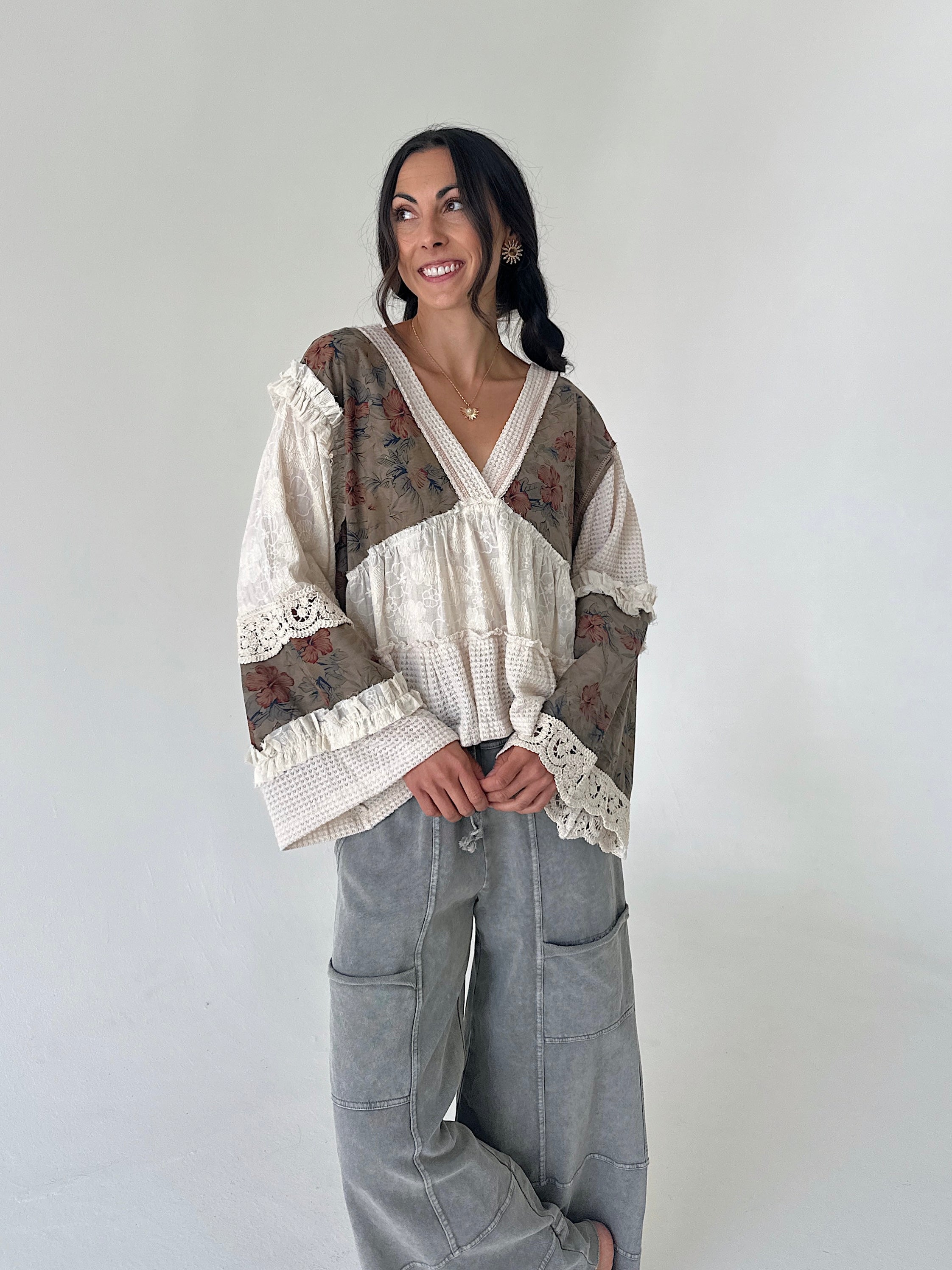 A woman modeling a bohemian-style patchwork blouse with floral and textured panels, lace trim, and wide sleeves, paired with gray wide-leg cargo pants against a plain light gray background.