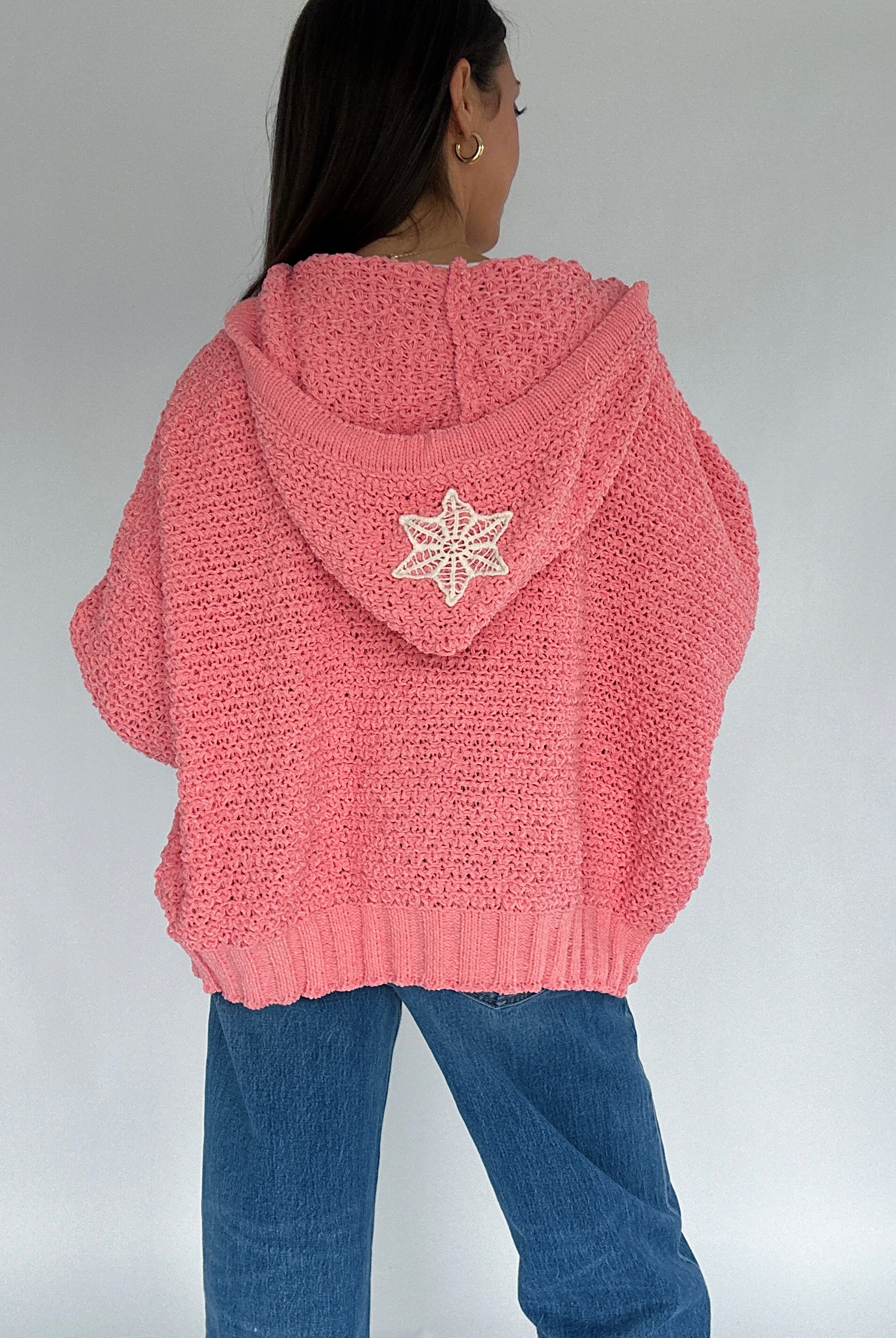 Person wearing a pink knitted hoodie with a crochet patch on the back against a plain background