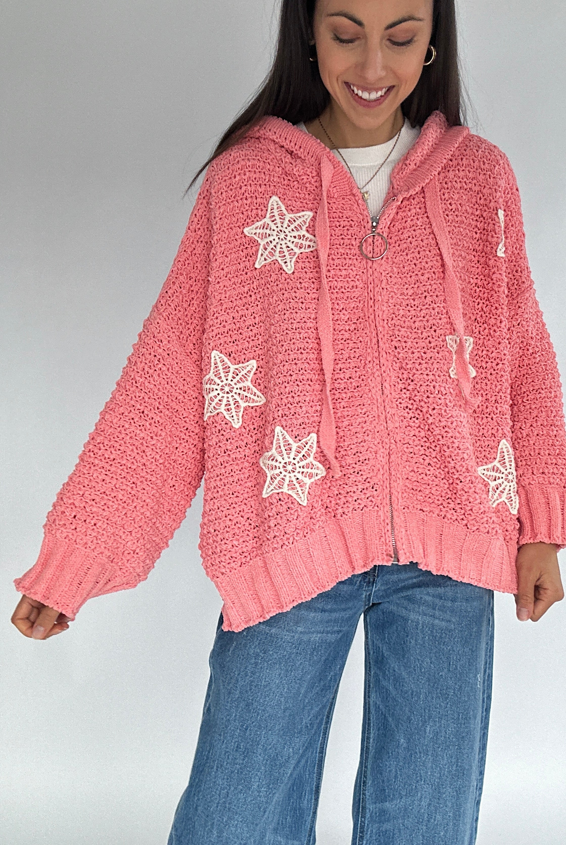Woman wearing a pink knitted zip up cardigan with crocket patterns on a plain background