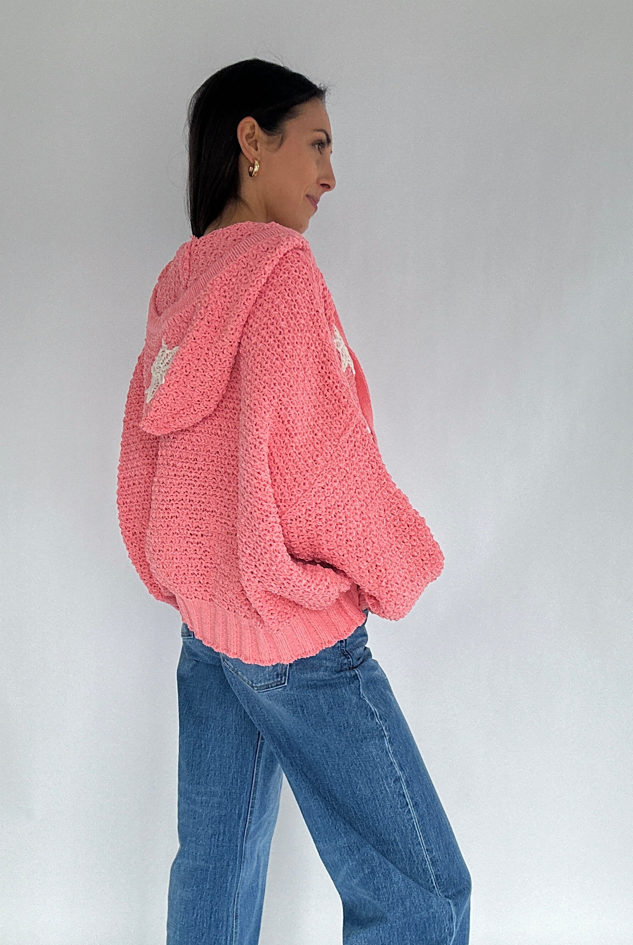 Person wearing a pink knitted hoodie sweater and blue jeans against a plain background