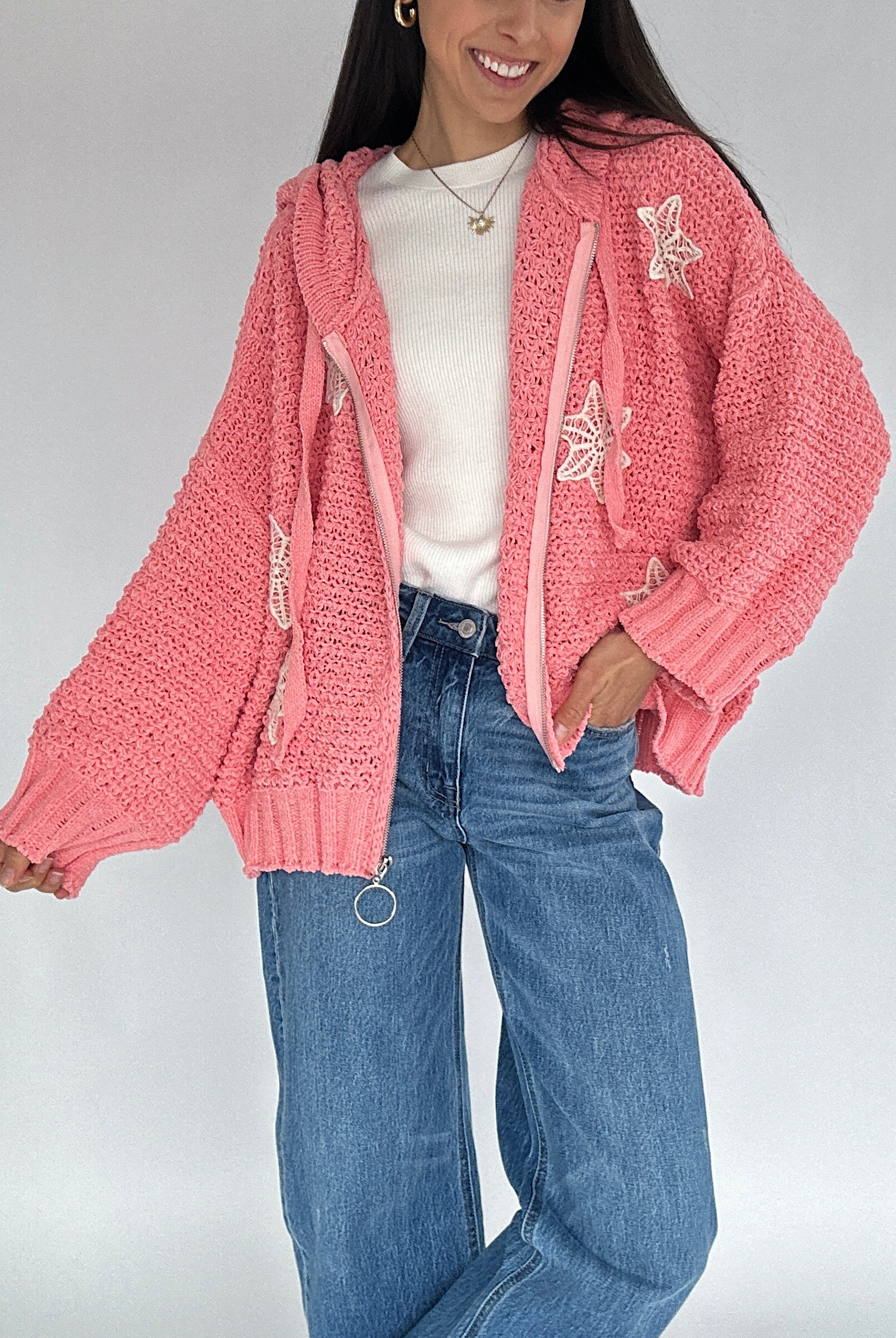 Person wearing a pink knit zip up cardigan with crocket designs, white shirt, and blue jeans on a plain background