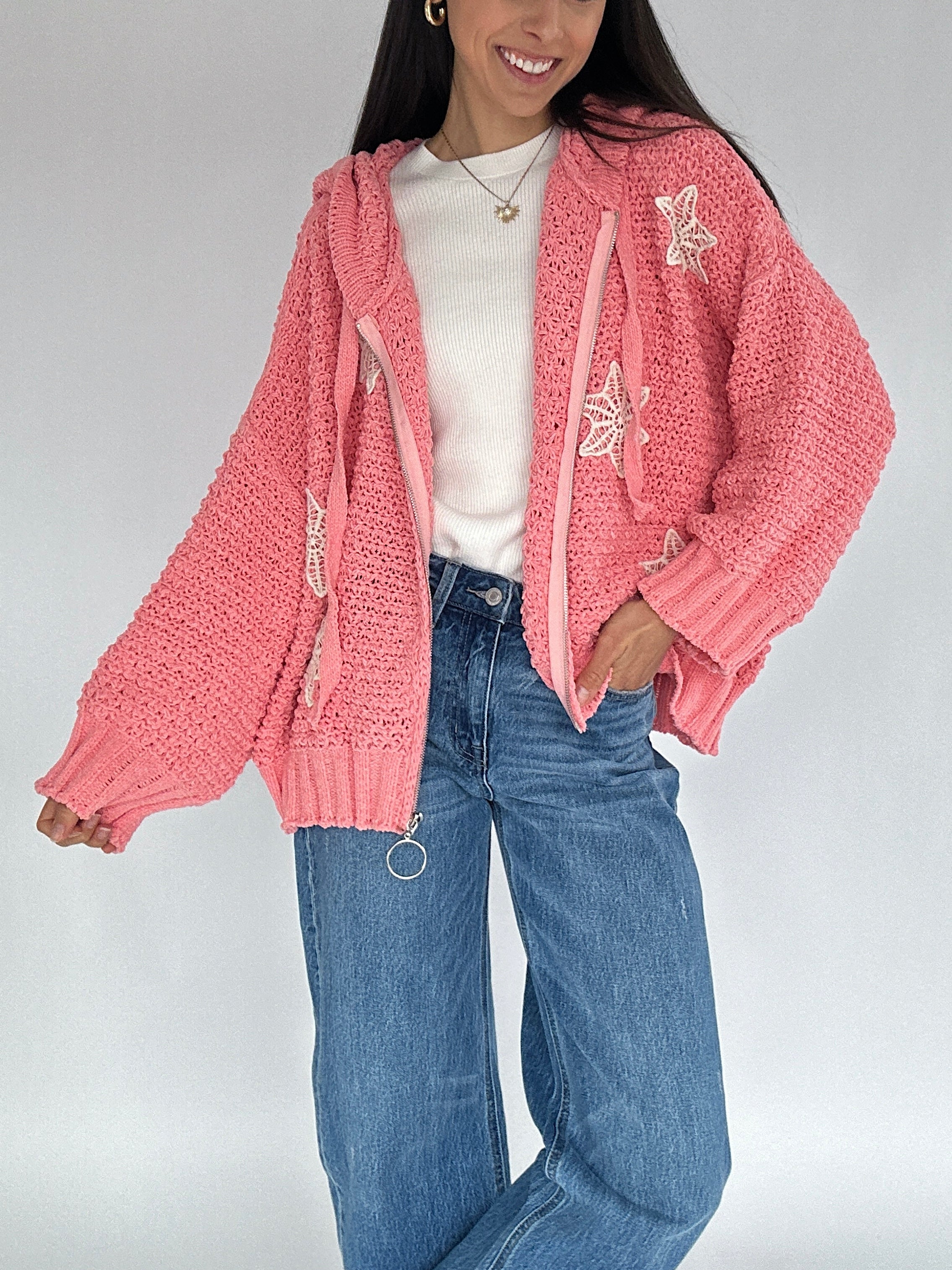 Person wearing a pink knit zip up cardigan with crocket designs, white shirt, and blue jeans on a plain background