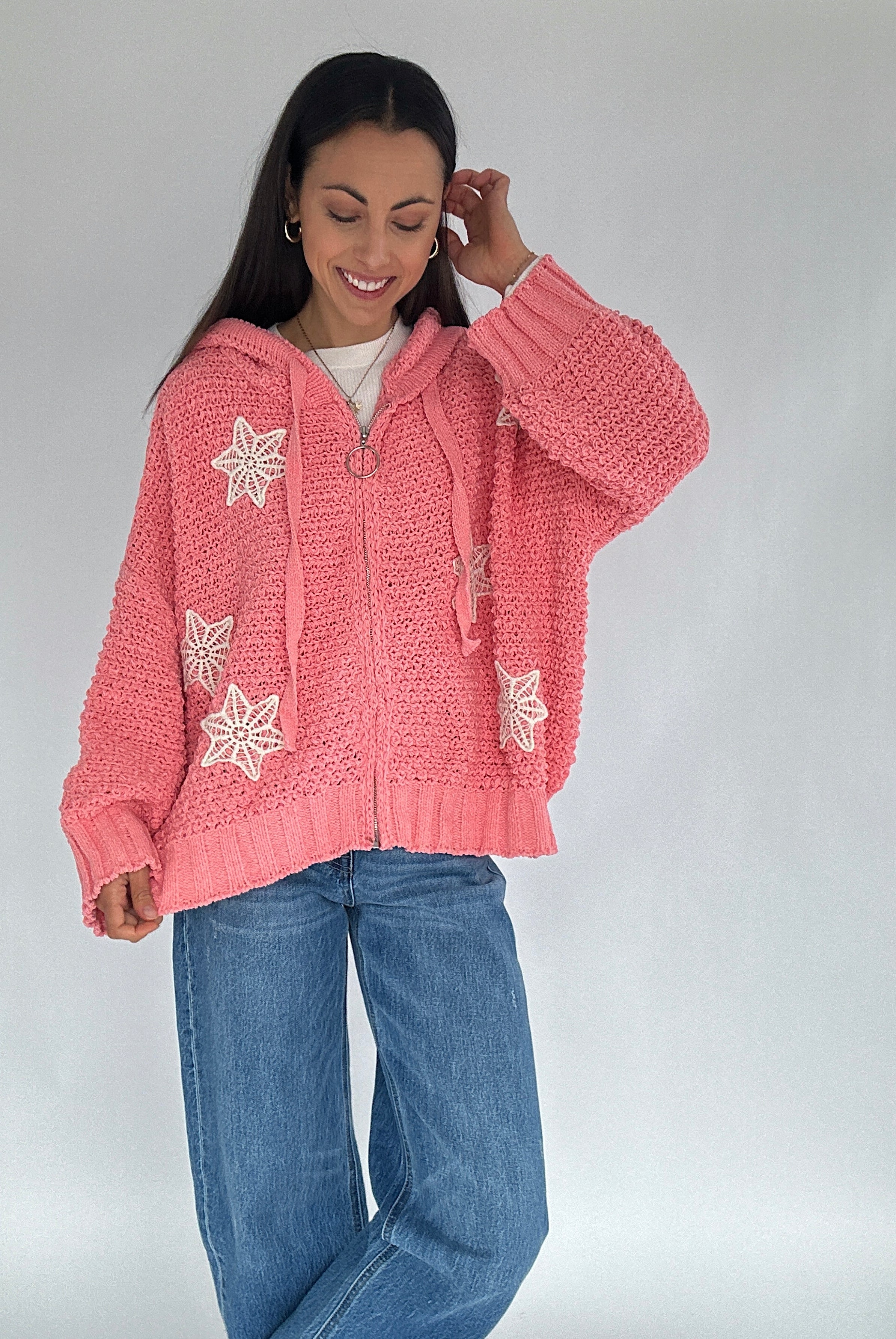 Woman wearing a pink knitted zip up hoodie sweater with white crochet patterns on a plain background