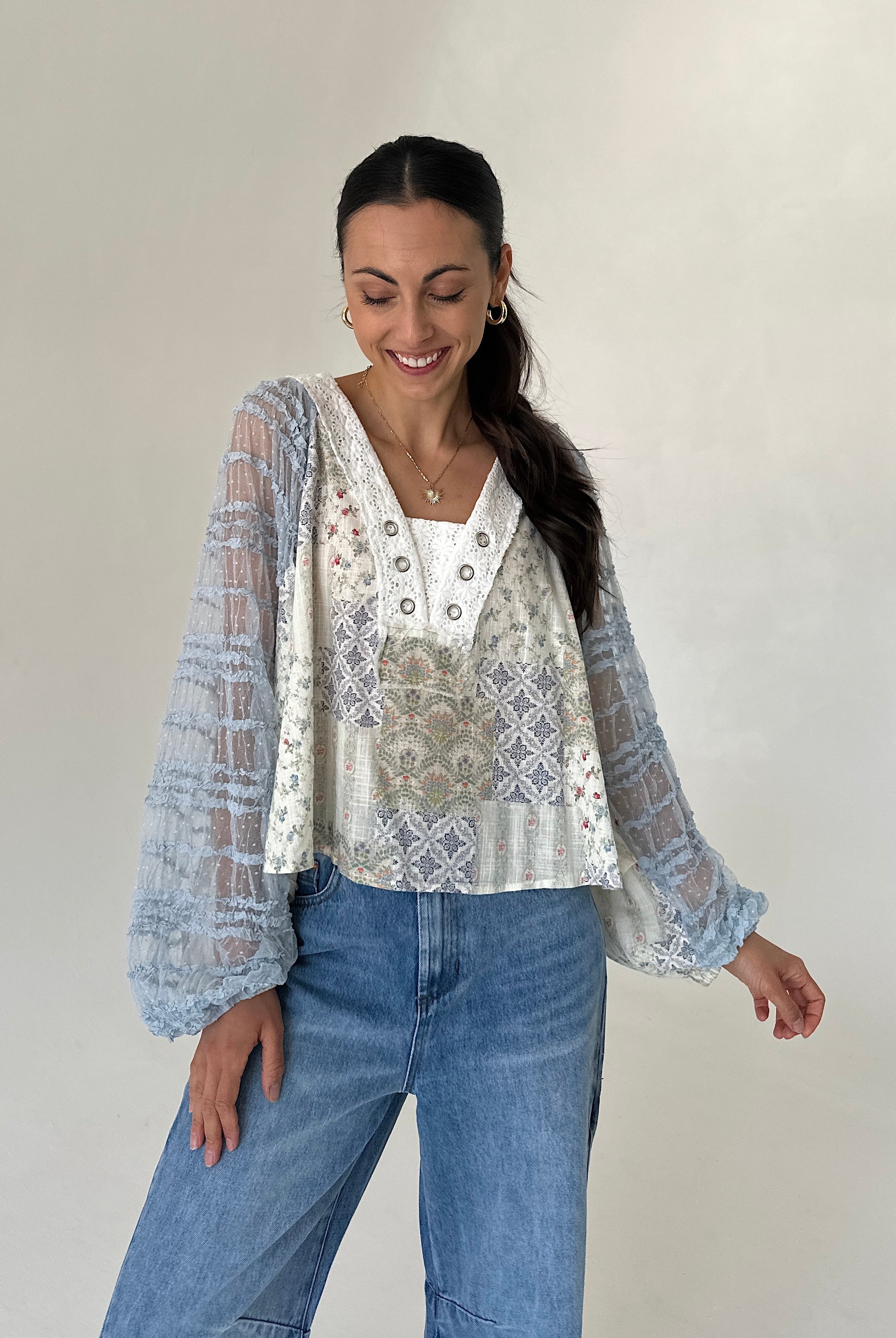 A woman modeling a printed blouse with sheer light blue patterned long sleeves, paired with blue jeans, against a plain light gray studio background.