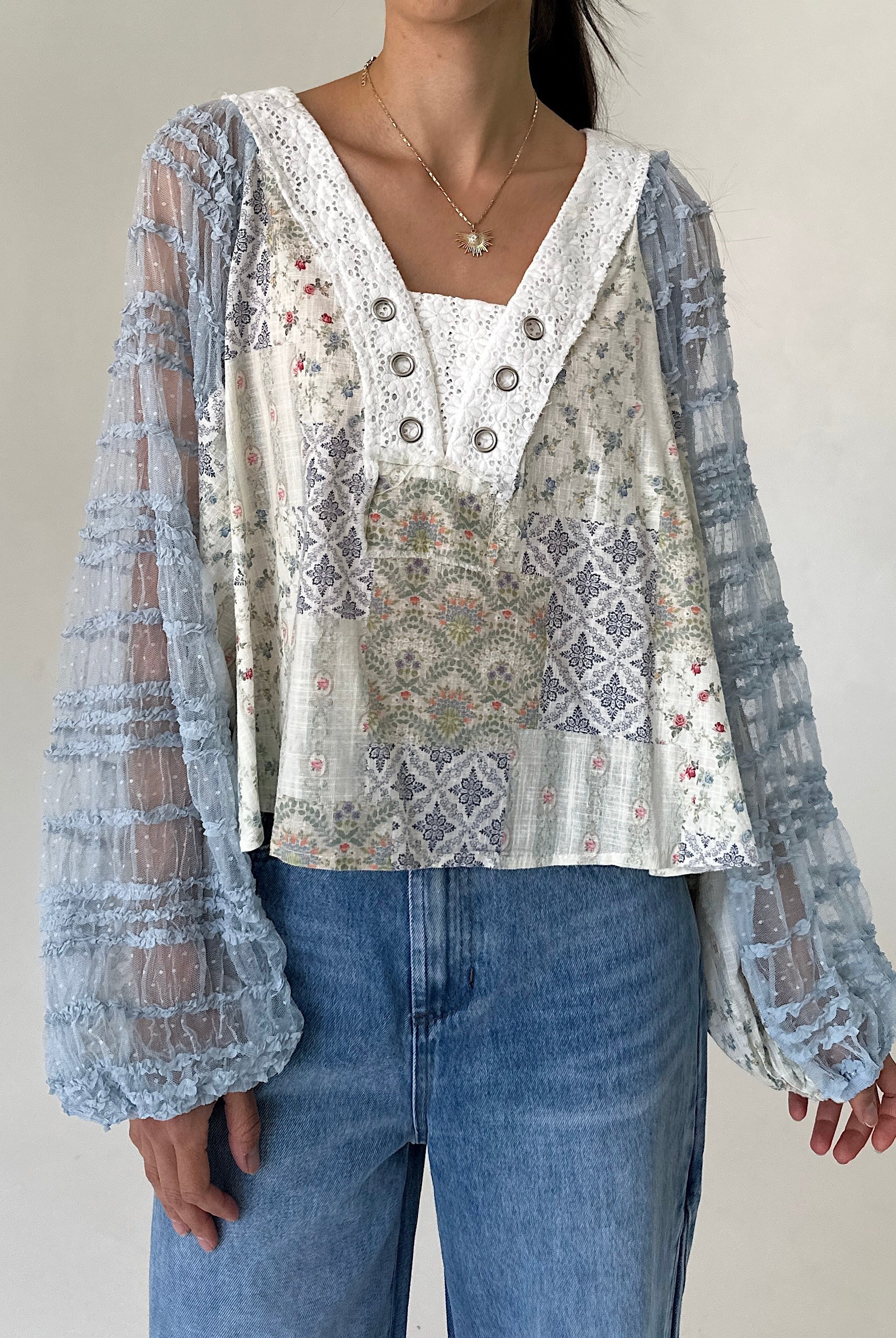 A woman wearing a bohemian-style patchwork blouse with sheer light blue lace long sleeves and a cream-colored embroidered bodice, paired with blue jeans.