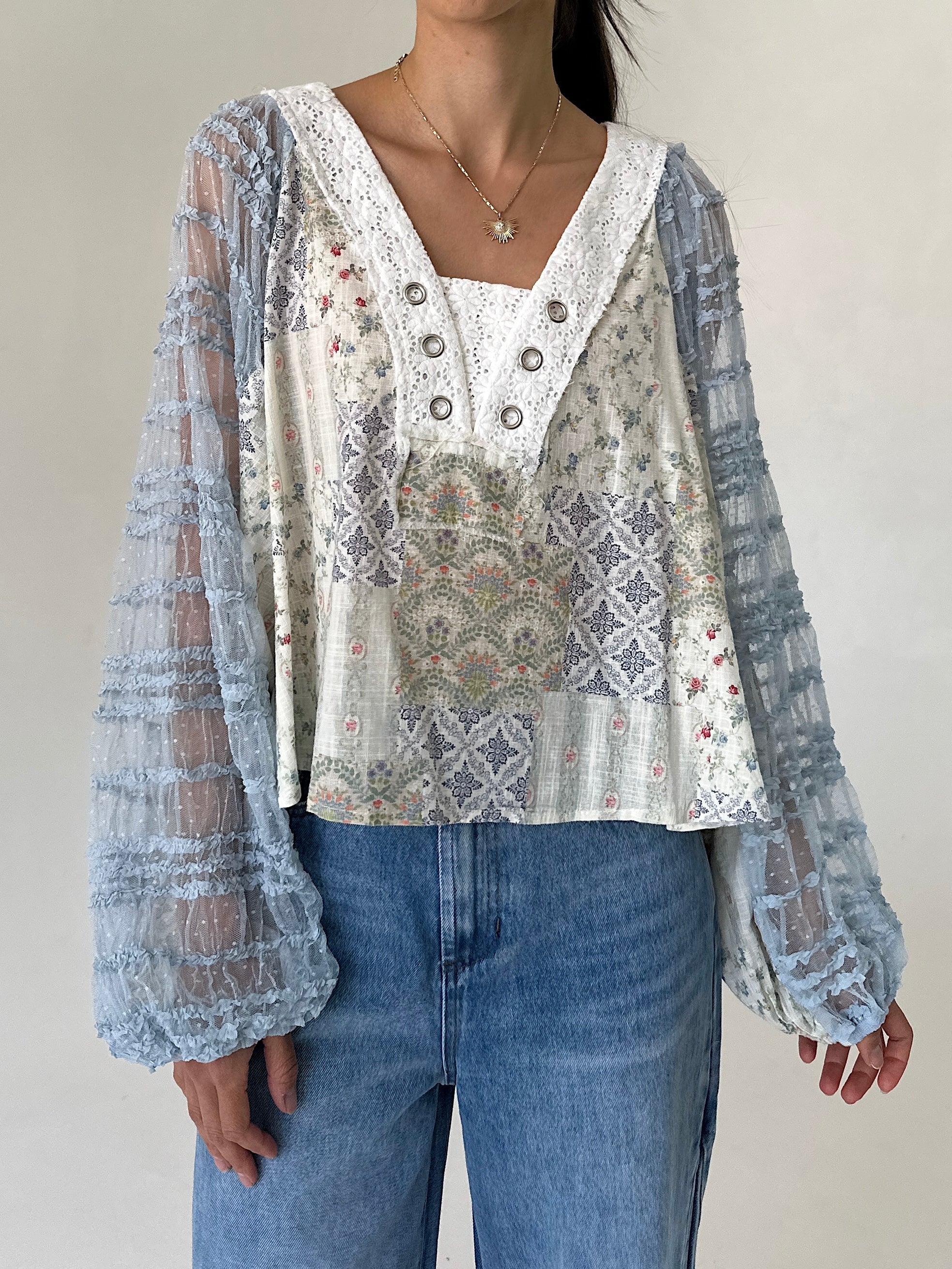 A woman wearing a bohemian-style patchwork blouse with sheer light blue lace long sleeves and a cream-colored embroidered bodice, paired with blue jeans.