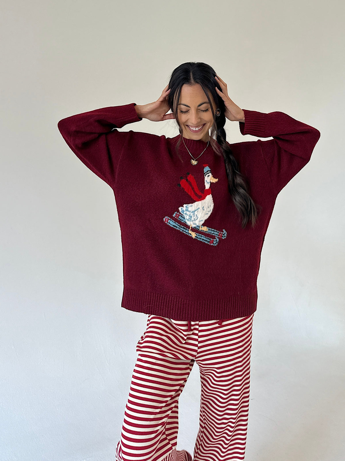 A woman wearing a burgundy knit sweater with a beaded goose graphic and red-and-white striped pants, posing against a plain off-white wall.