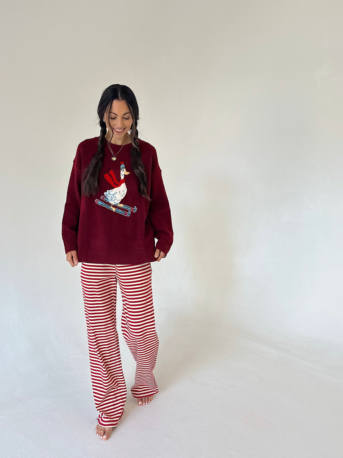 A woman modeling a cozy holiday outfit consisting of a deep burgundy long-sleeve top with a red goose graphic and matching red-and-white striped pants, standing against a plain off-white studio background.