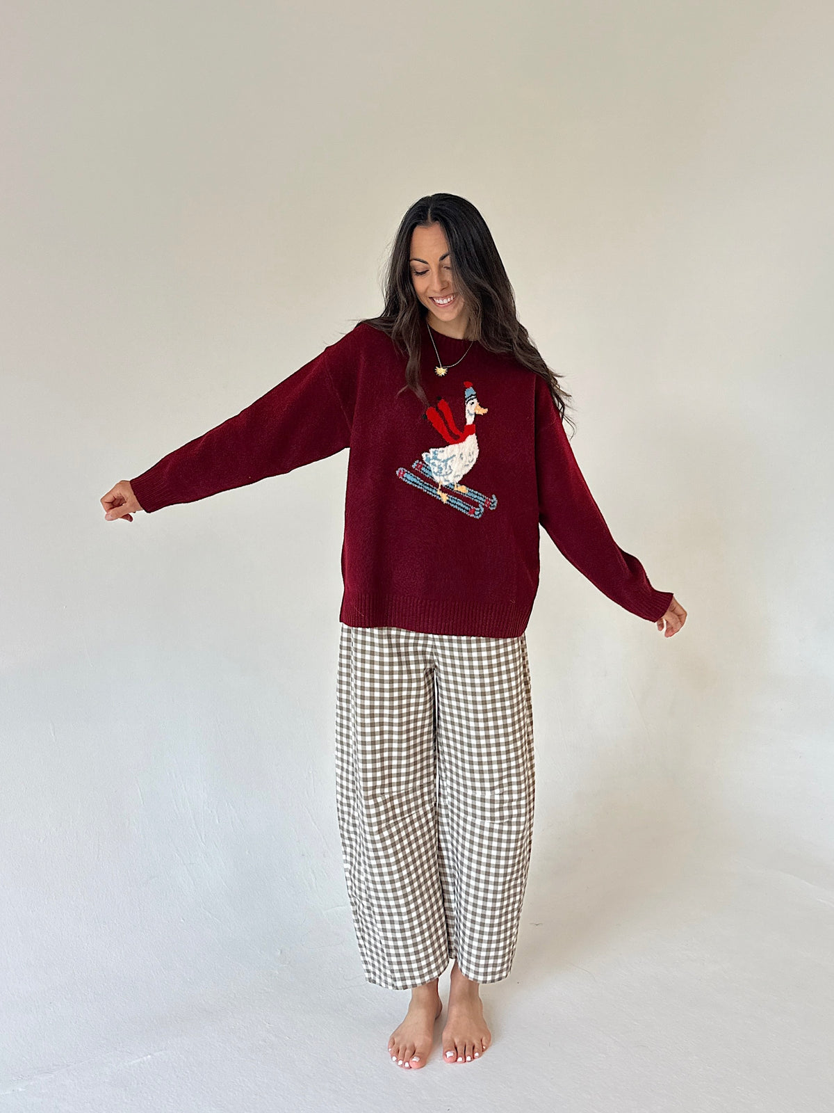 A woman stands barefoot against a plain off-white studio background, wearing a burgundy knit sweater with a white goose graphic on the front and beige-and-white gingham pajama pants.