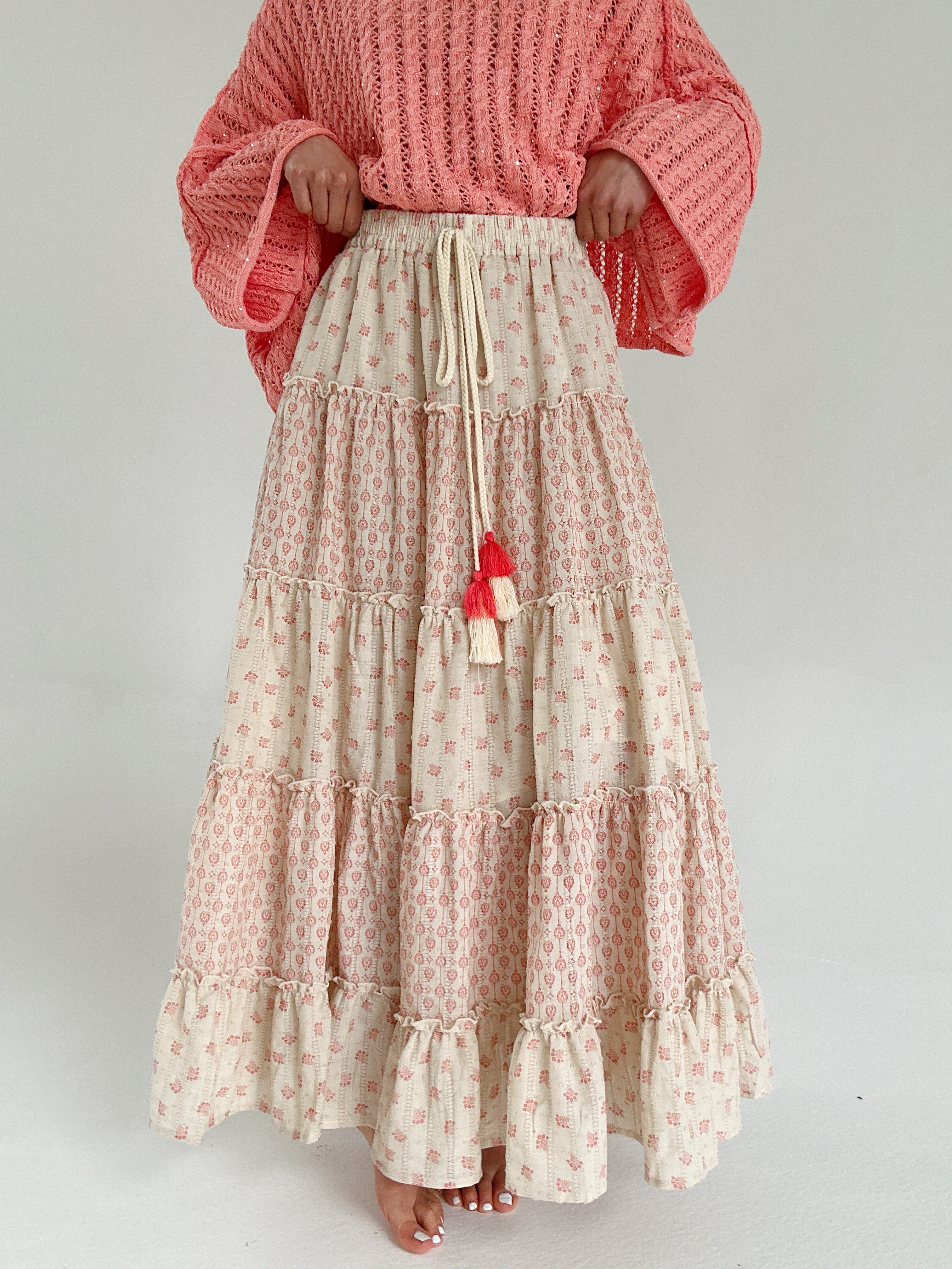 Person wearing a coral patterned sweater and a long, tiered skirt with a drawstring on a plain background