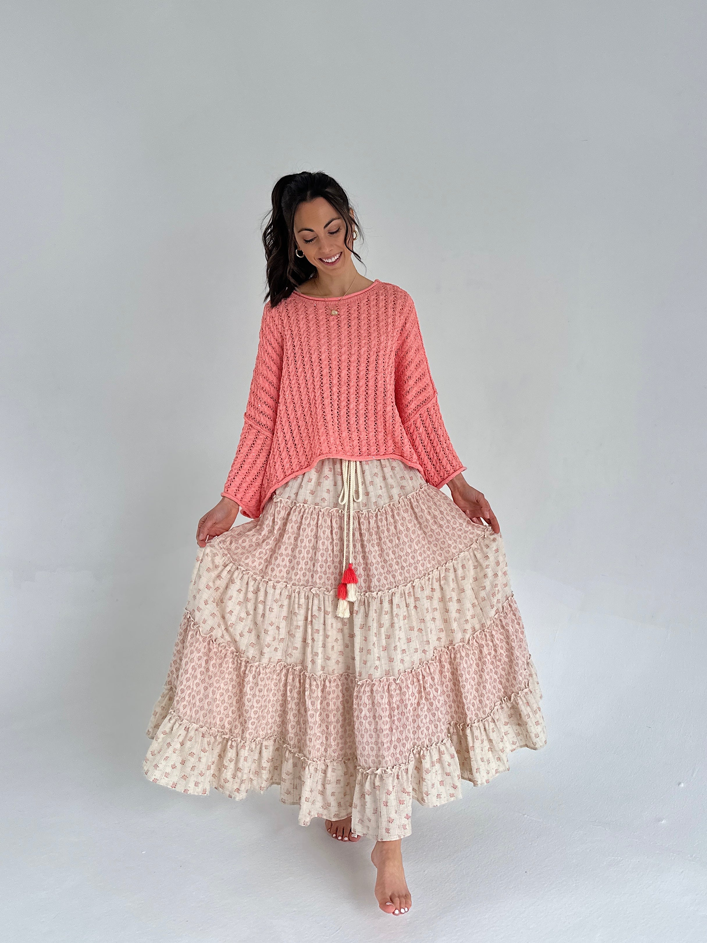 Woman wearing a coral sweater and a long, patterned skirt on a plain background