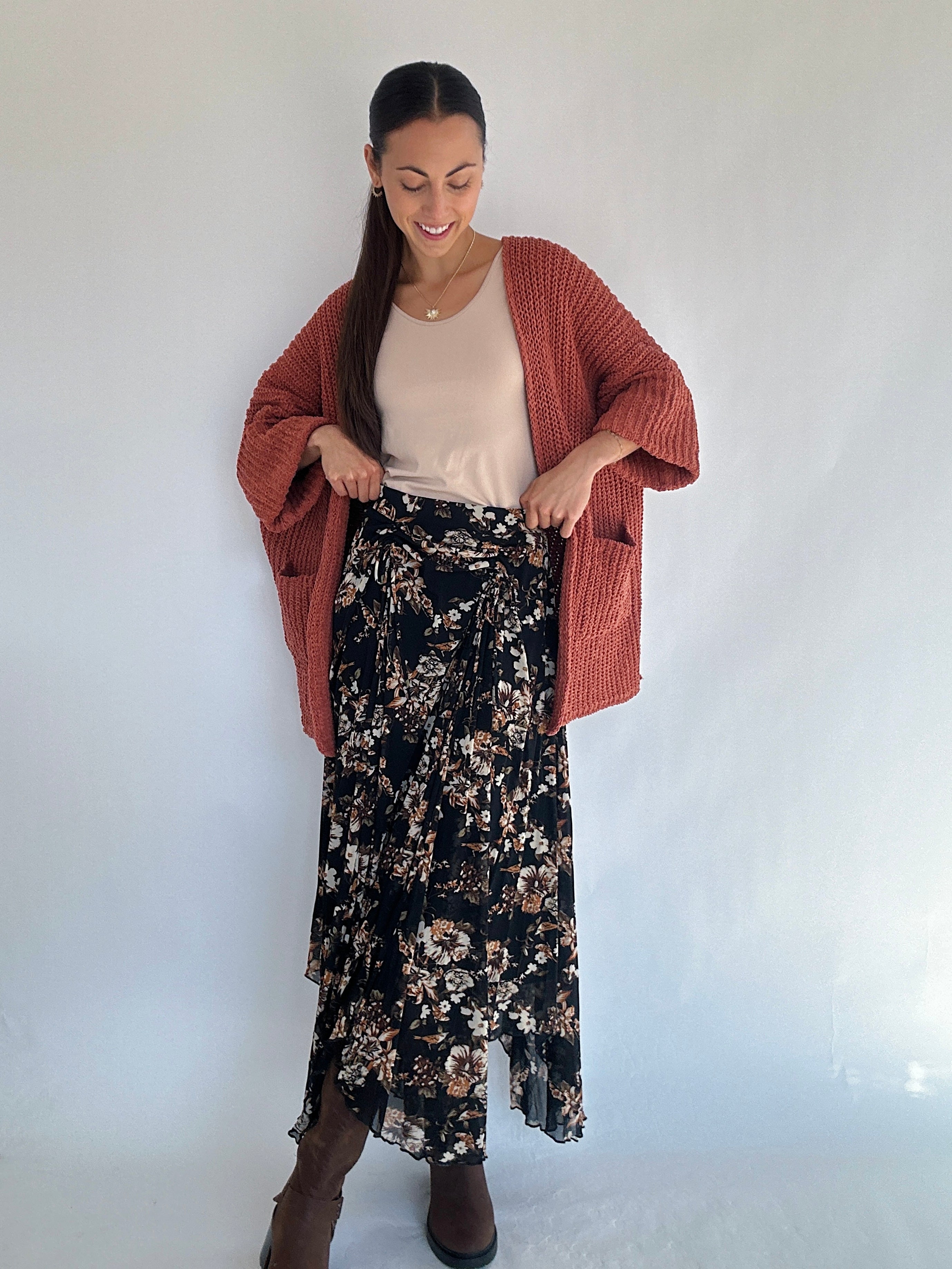 Woman wearing a rust-colored cardigan, beige top, and black floral skirt against a white background