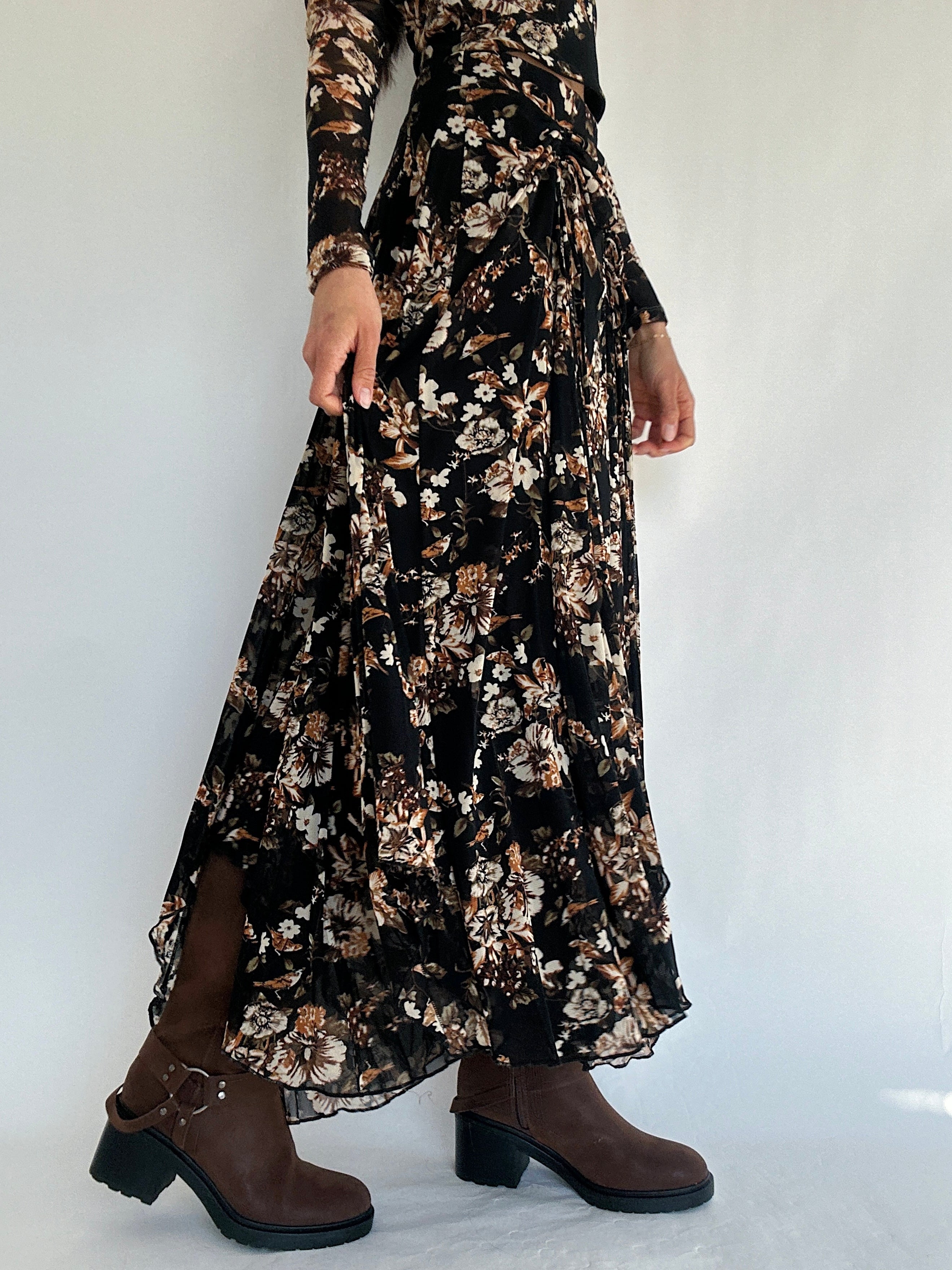 Person wearing a long black floral mesh skirt with brown boots on a white background showing side view