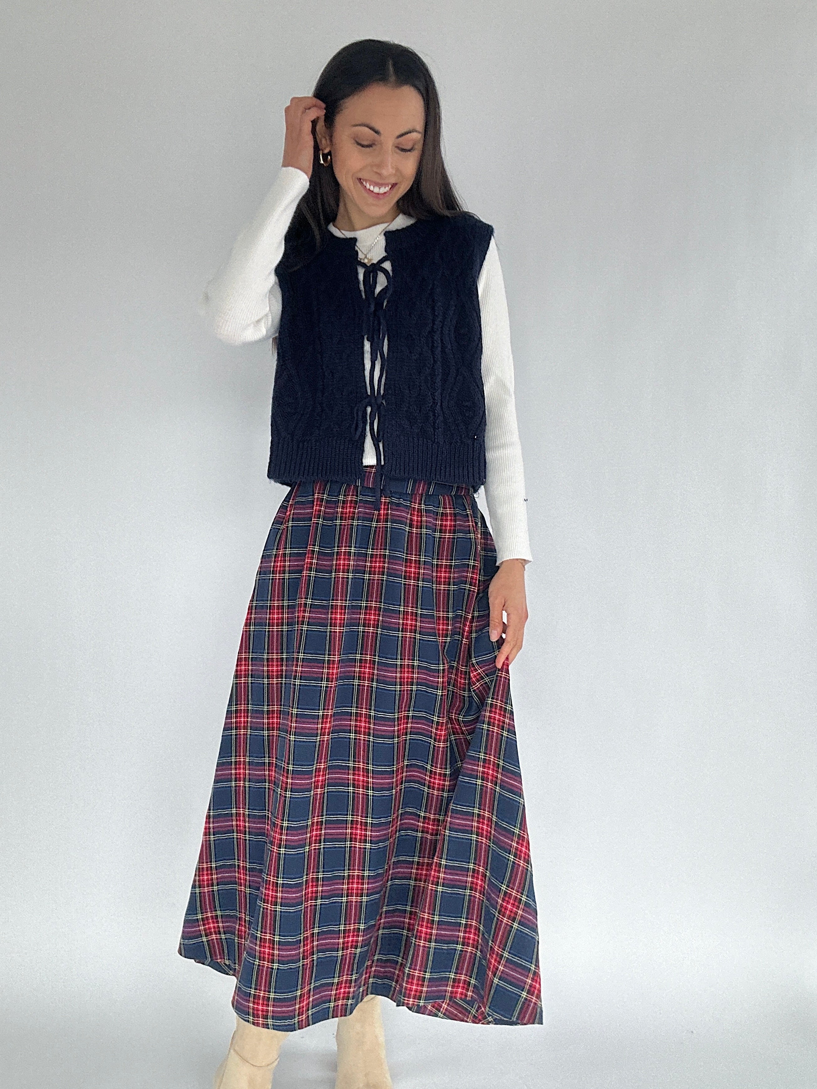 Woman wearing a navy vest over a white shirt with a red plaid skirt on a plain background