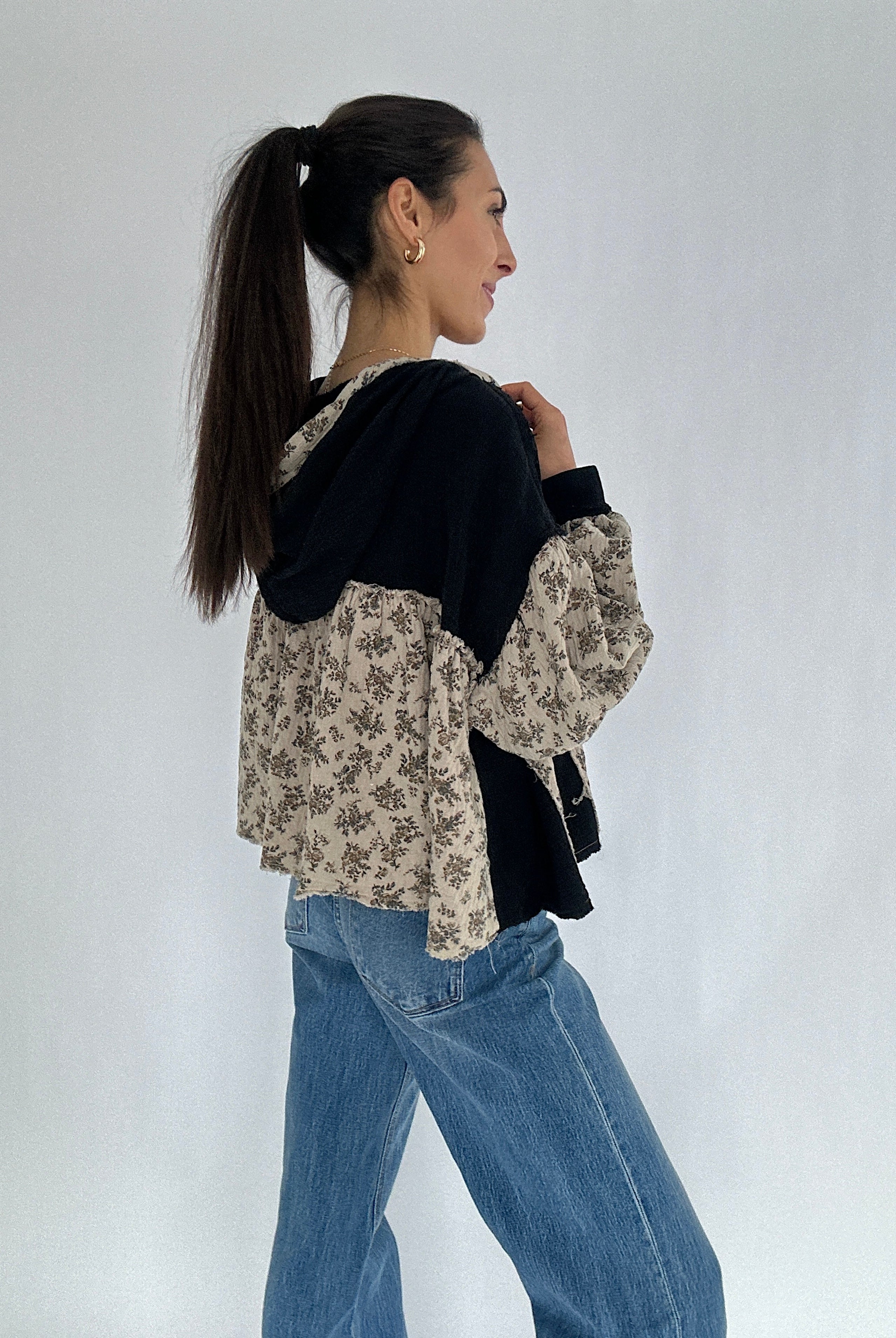 Woman wearing a black and floral hooded top and blue jeans on a white background showing side angle