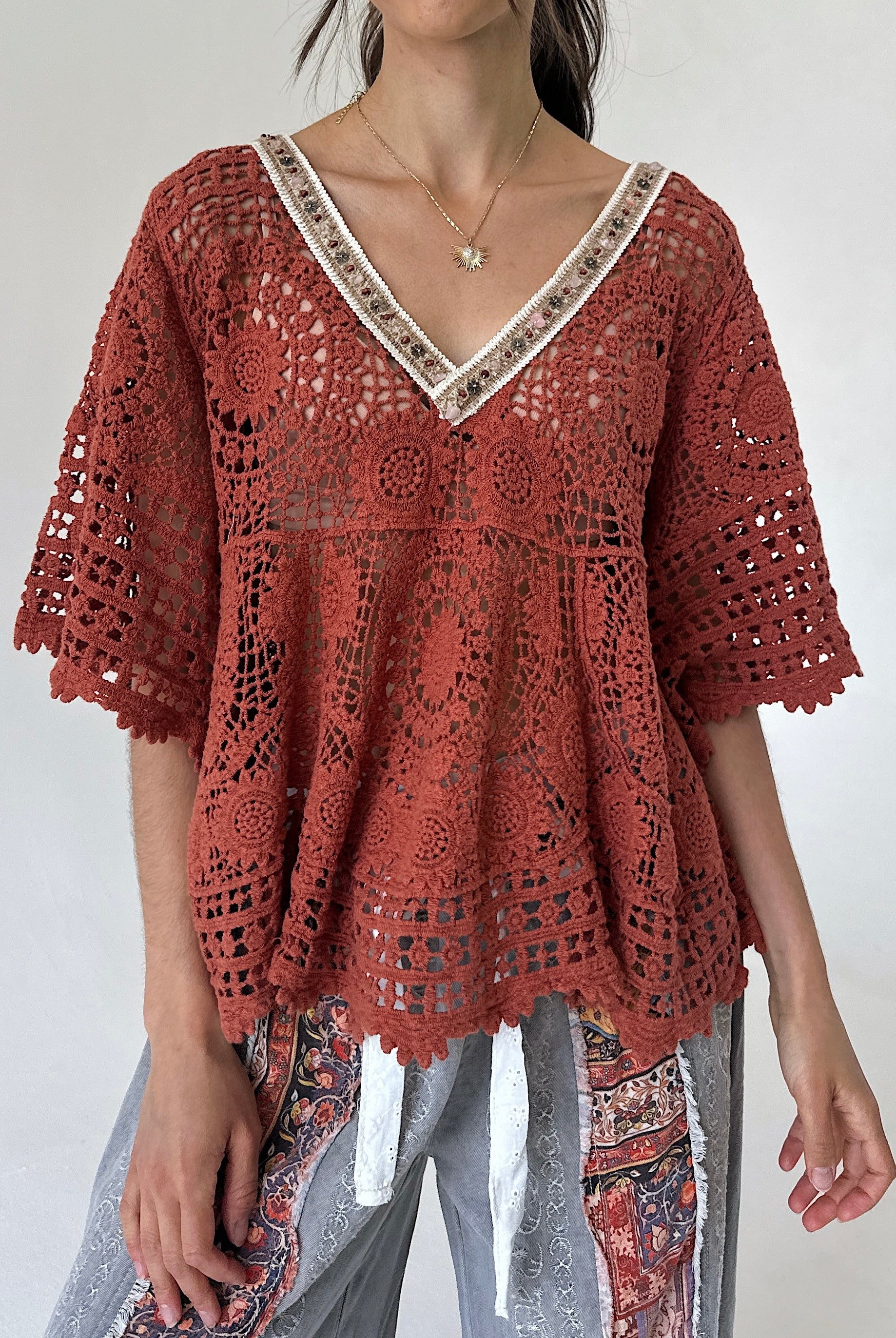 A woman wearing a rust-colored crochet lace top with a V-neck and patterned gray pants, photographed against a plain light background.