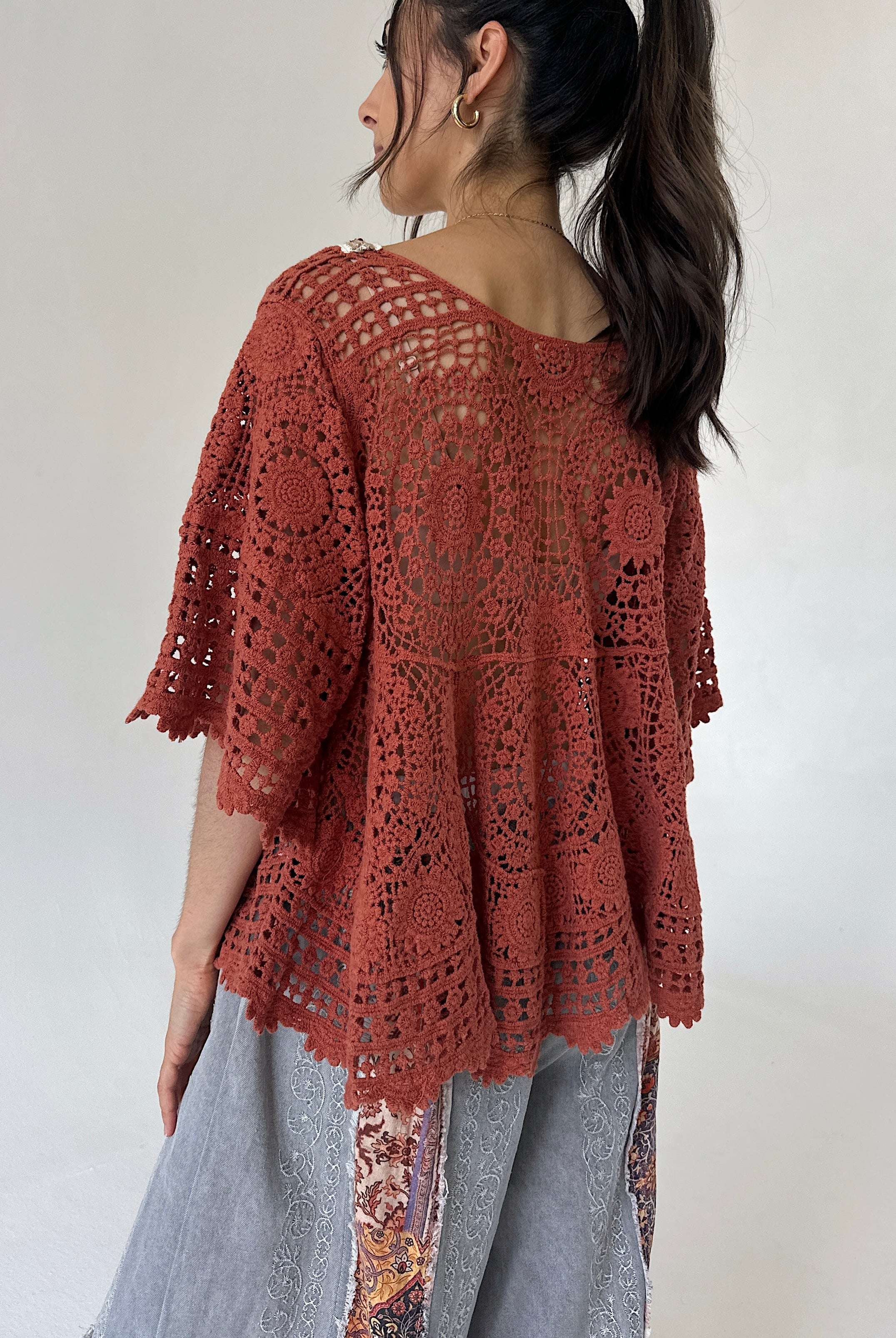 A woman shown from the back wearing a rust-colored, open-knit crochet style top over light gray patterned pants, set against a plain light gray background.