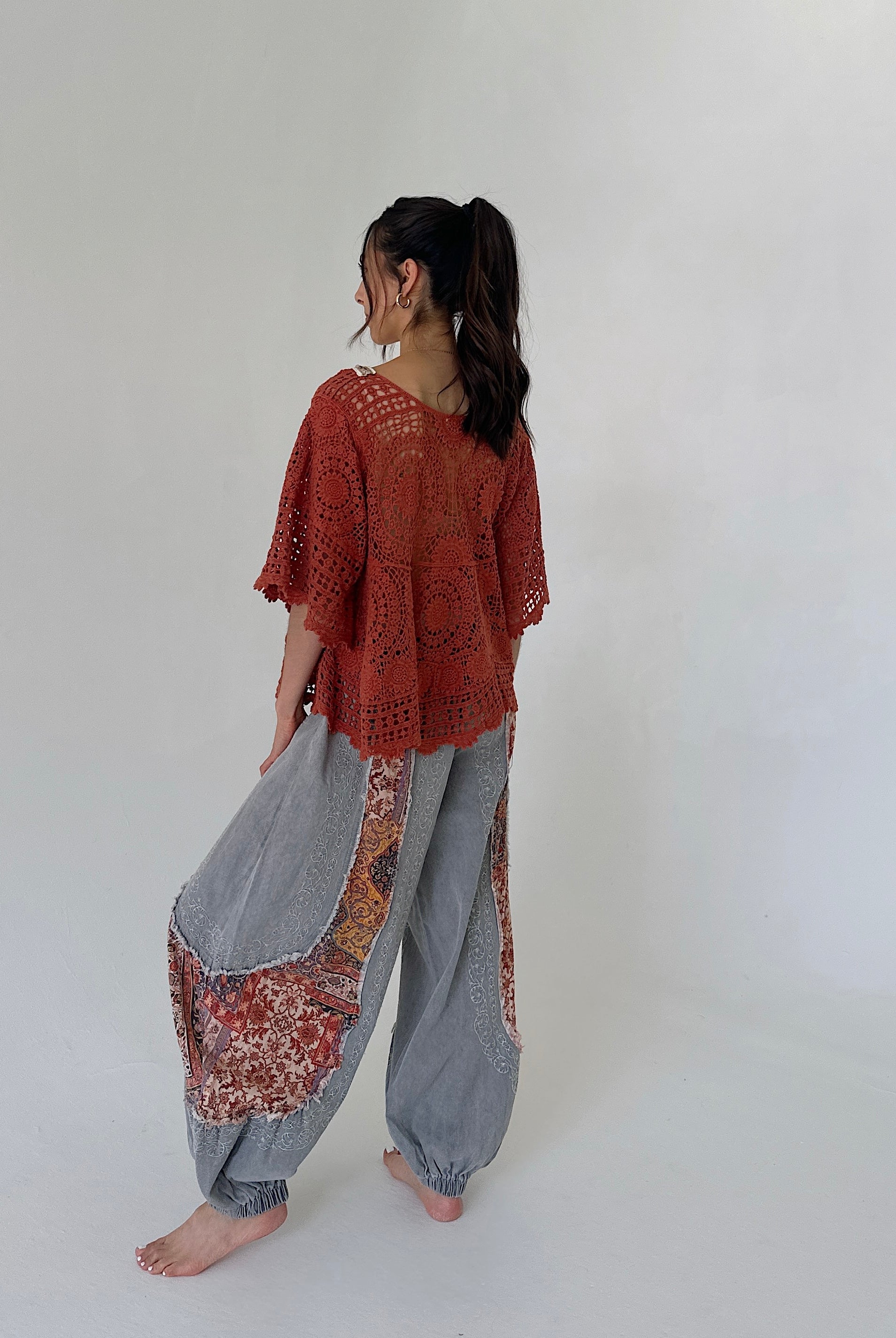 A woman stands barefoot against a plain light gray studio background, shown from the back. She is wearing a loose rust-orange crochet short-sleeve top and wide-leg gray harem-style pants with rust-orange patterned patch panels near the lower legs.