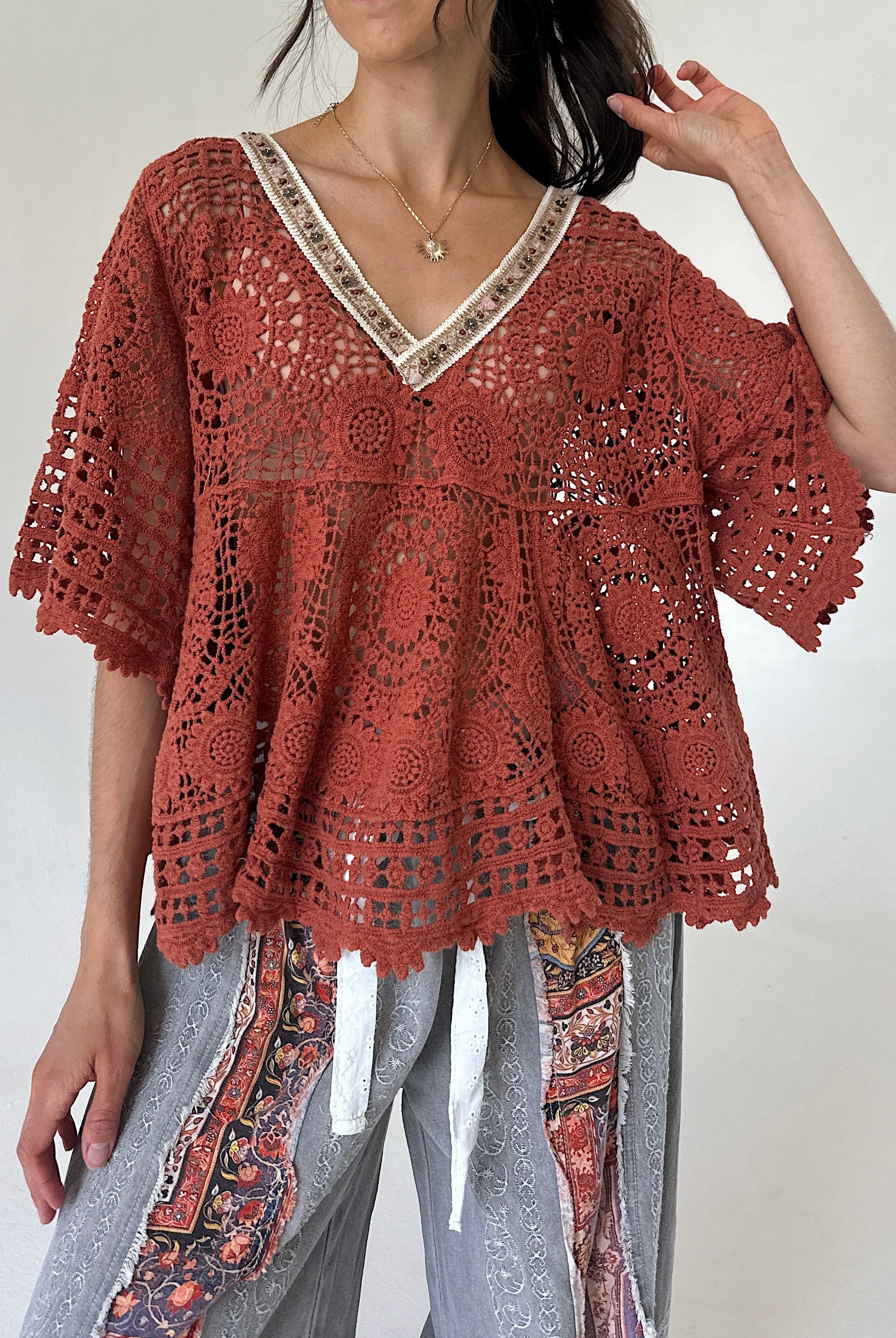 A woman wearing a rust-colored crochet lace top with a V-neck and short sleeves, paired with patterned gray pants, against a plain light background.