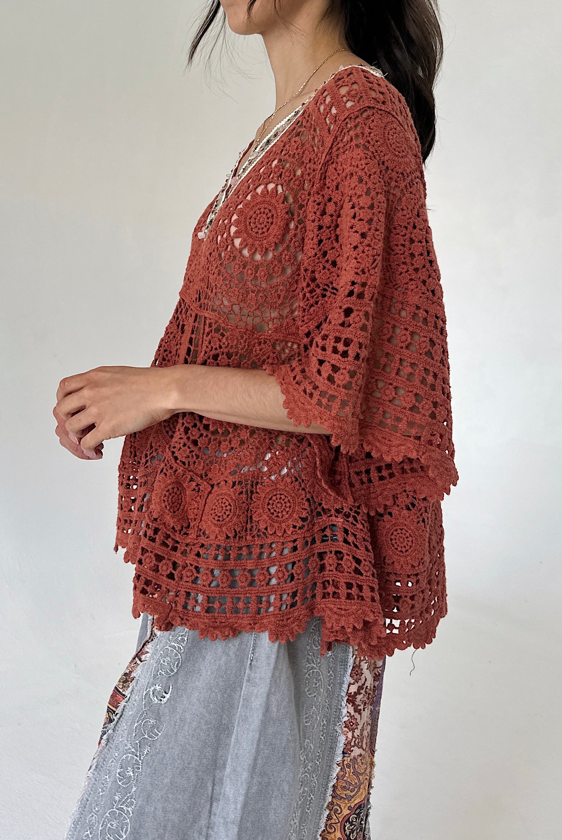 A woman wearing a rust-colored crochet poncho top with wide three-quarter sleeves and a scalloped hem, paired with light gray patterned pants, shown from a side profile against a plain light background.