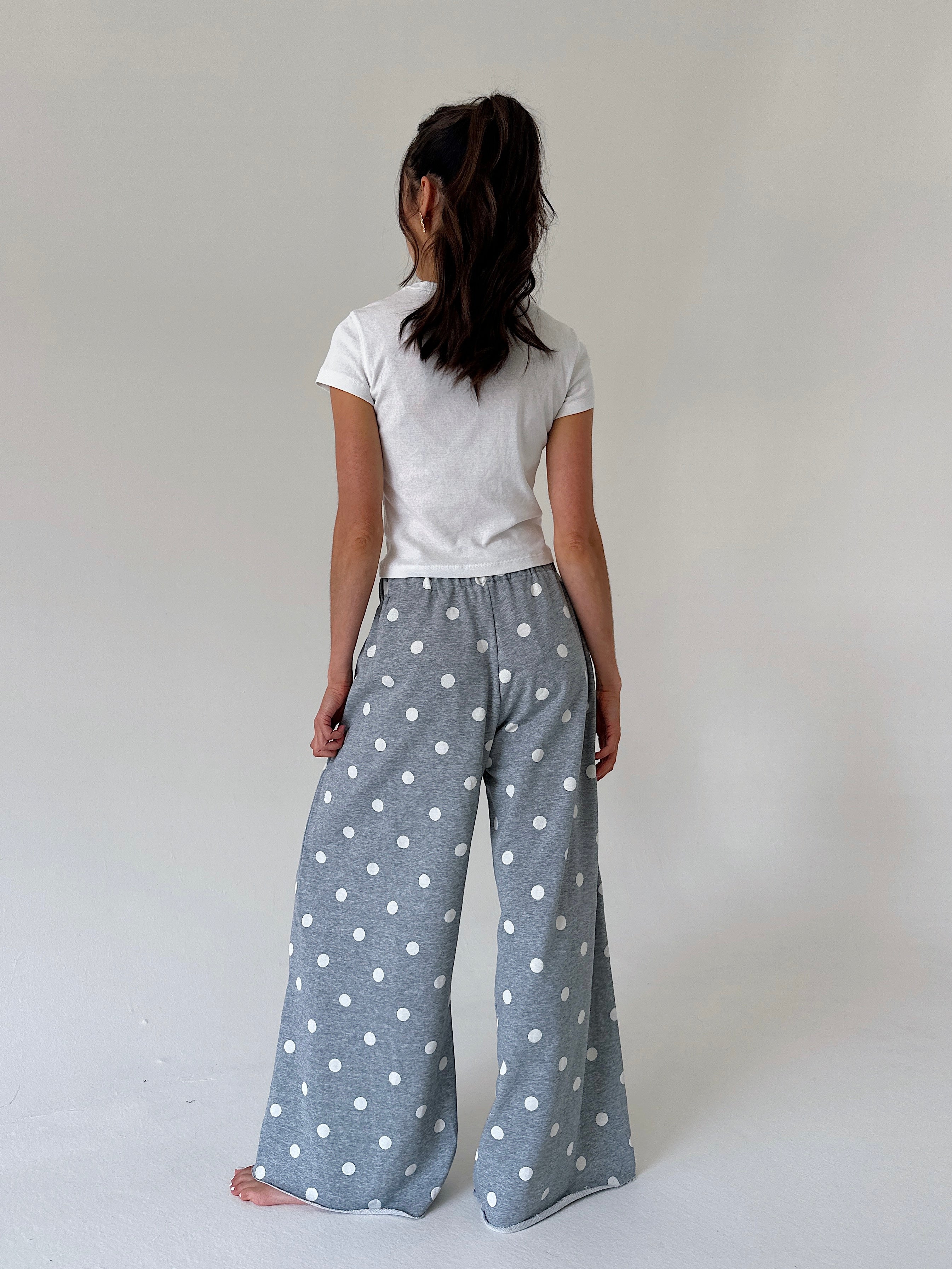 A woman viewed from behind wearing wide-leg gray pants with a white polka dot pattern, paired with a white t-shirt, standing against a plain light gray studio background.