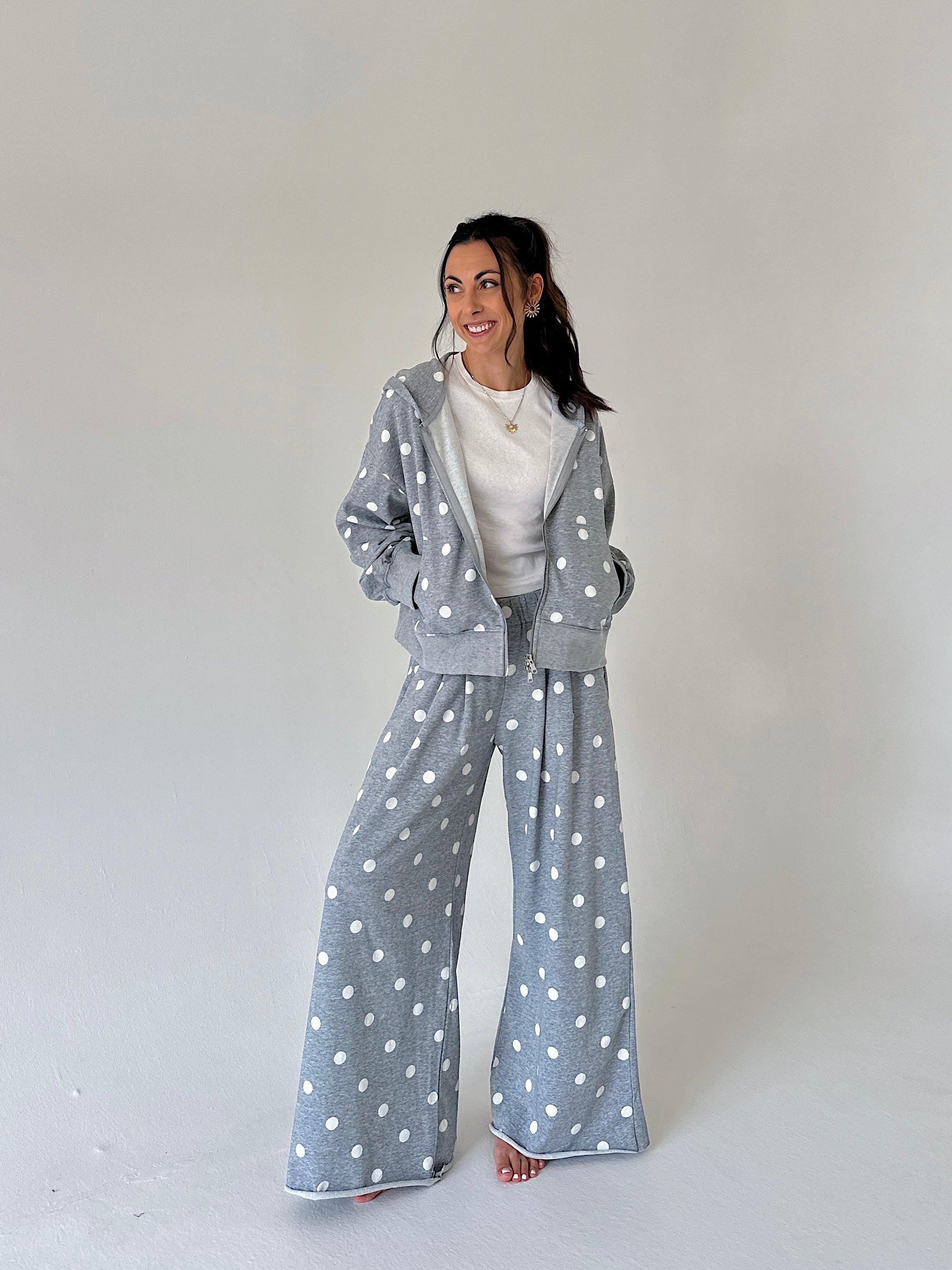 A woman modeling a matching gray two-piece loungewear set with white polka dots, consisting of a zip-up hoodie and wide-leg pants, styled over a white t-shirt against a plain light gray studio background.