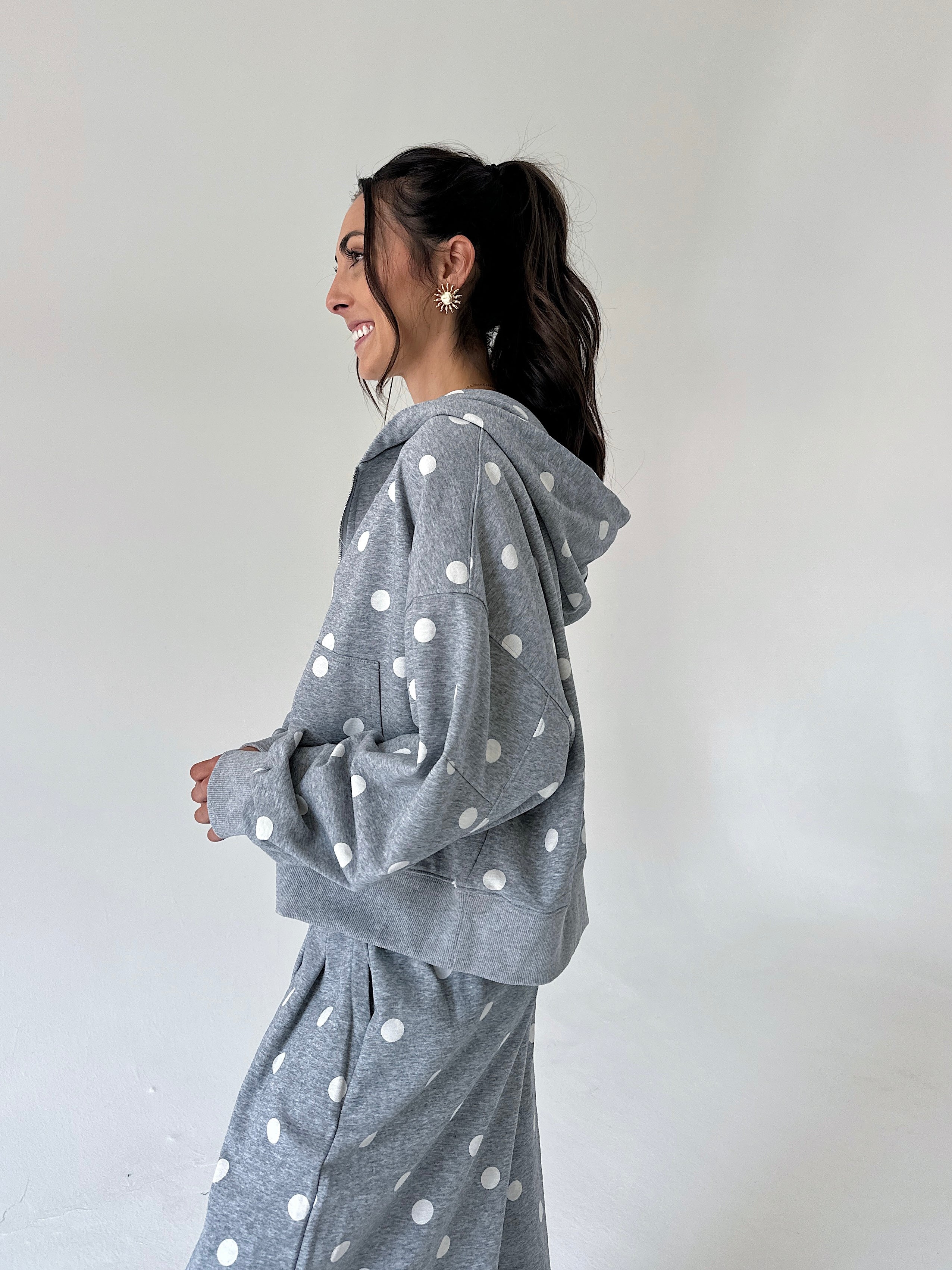 A woman wearing a gray hooded sweatshirt and matching sweatpants with white polka dots, shown in profile against a plain light gray studio background.