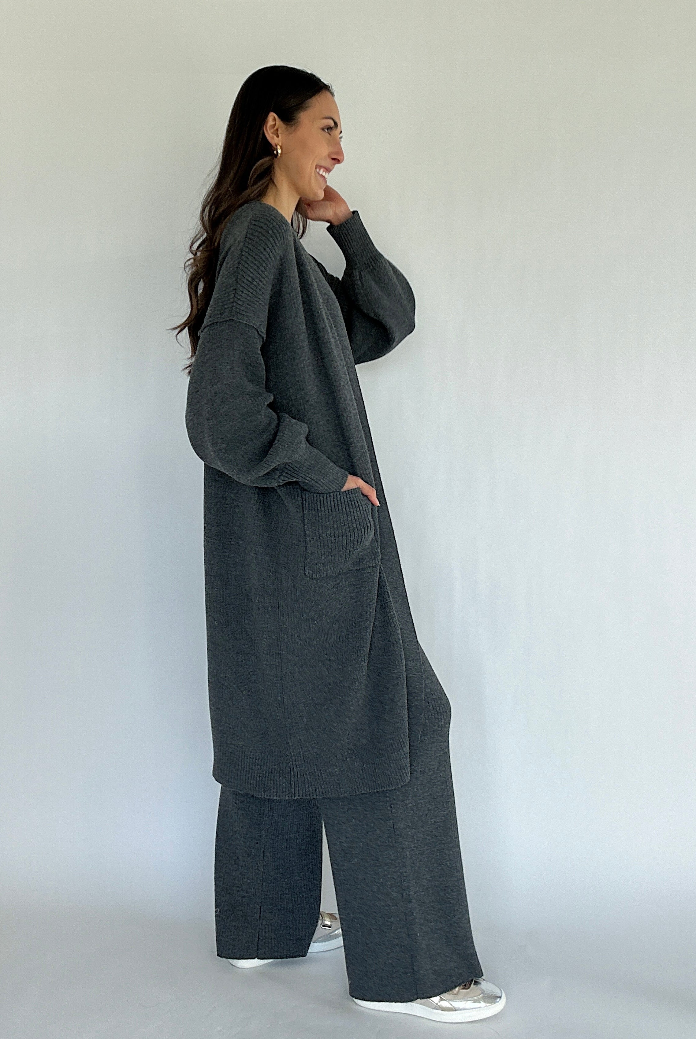Woman wearing a long dark gray cardigan and matching pants against a white background from side