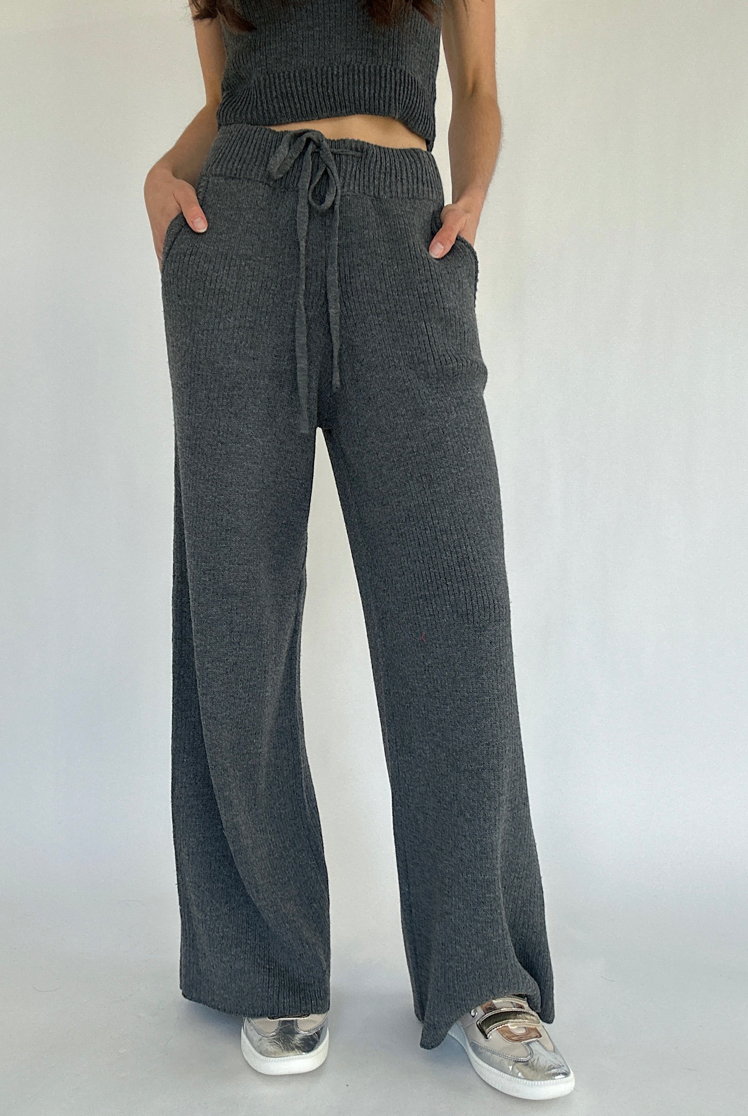Person wearing dark gray knit pants with a tied waist on a plain background with hands in pockets