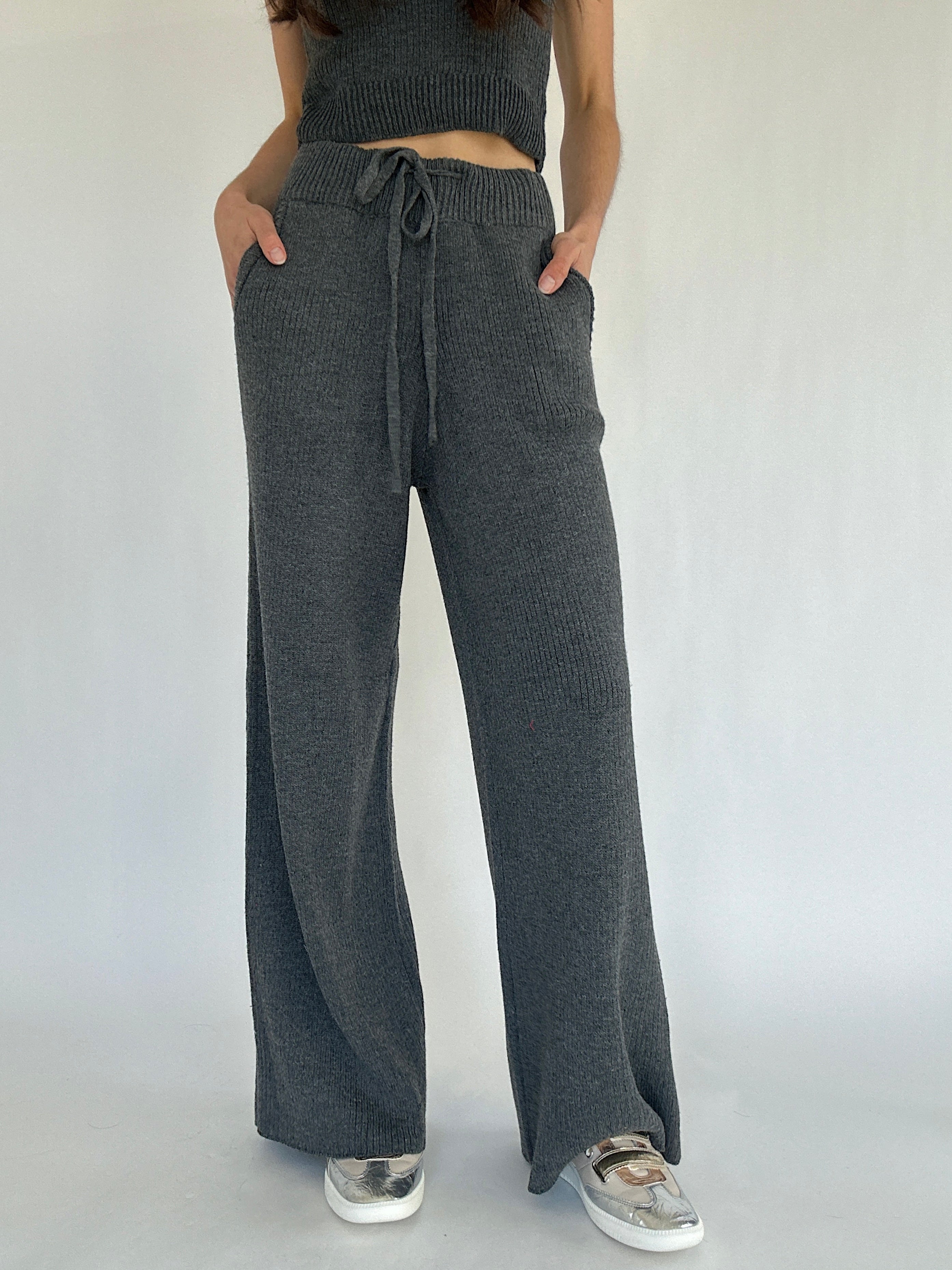 Person wearing dark gray knit pants with a tied waist on a plain background with hands in pockets