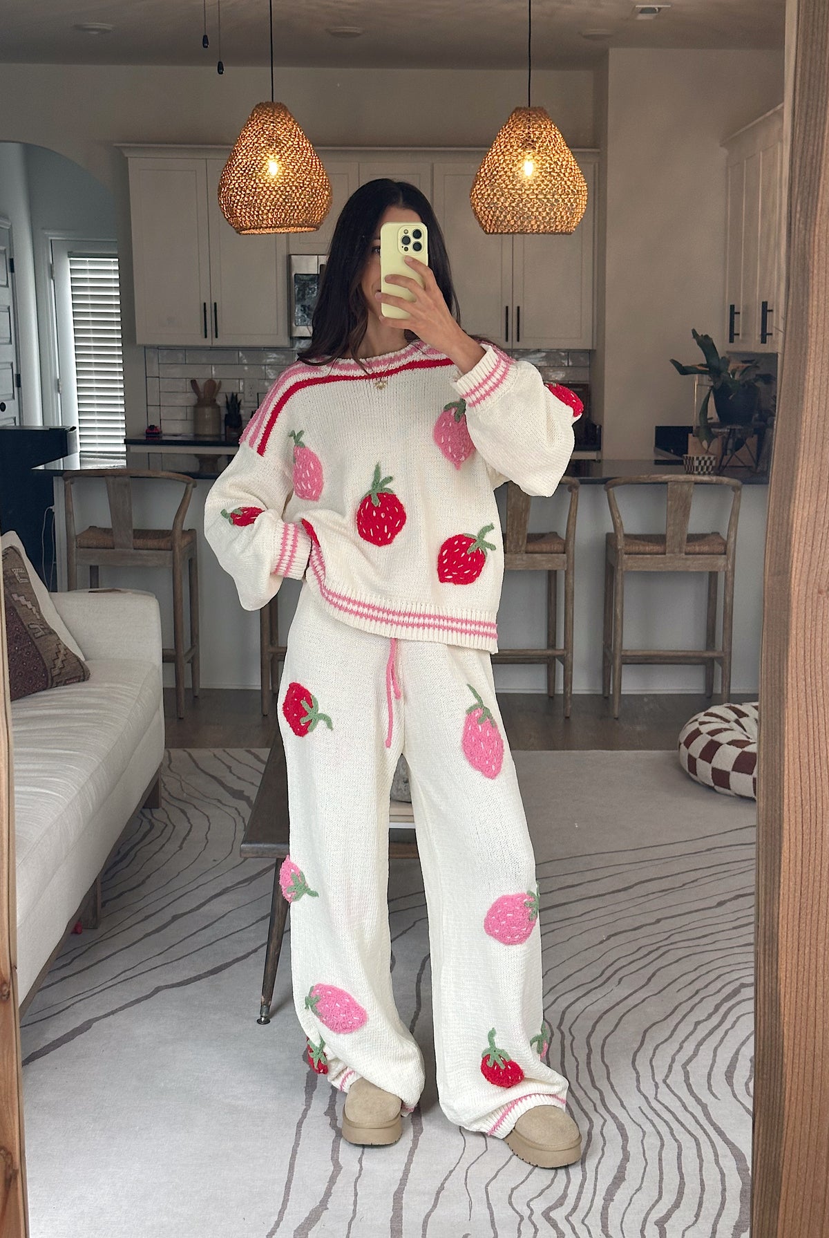 A woman takes a mirror selfie in a cozy room wearing a matching white two-piece set with pink and red strawberry prints. The set includes a long-sleeve top and wide-leg pants, styled with beige slippers.