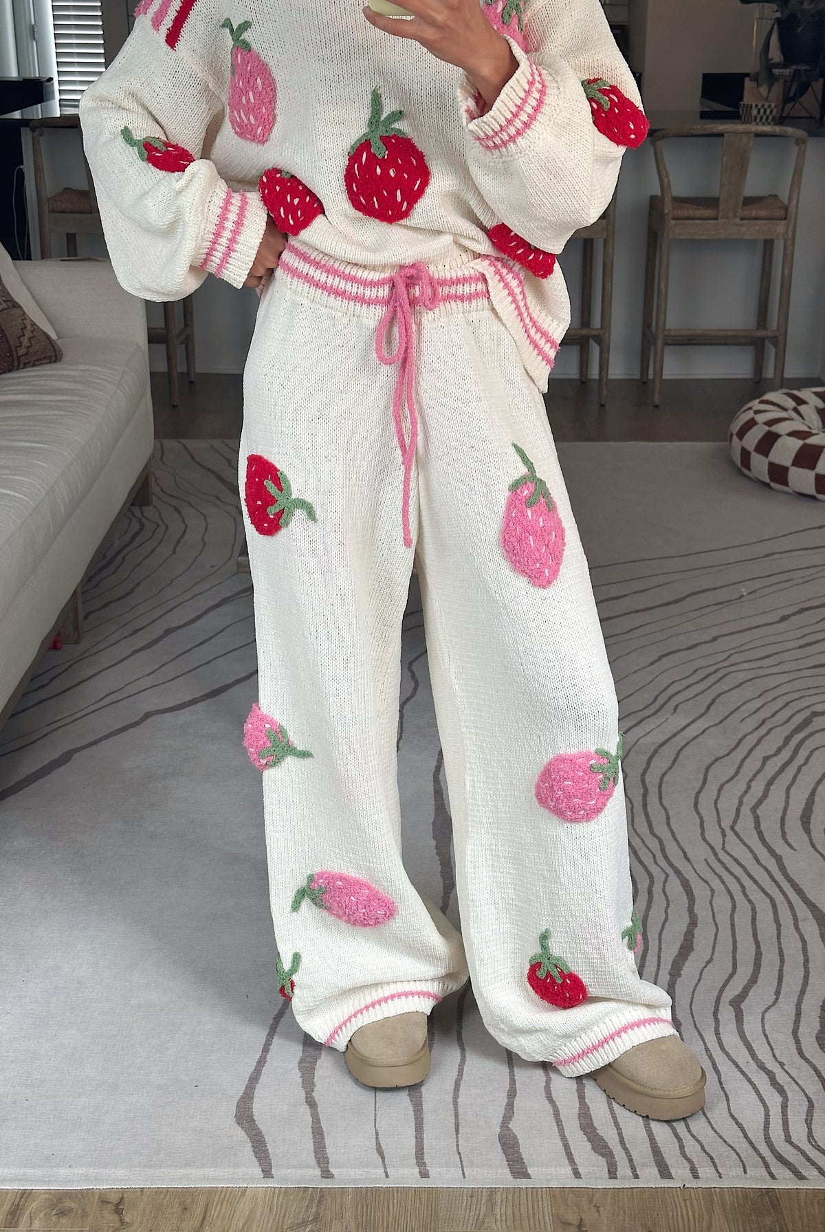 A person wearing a cozy knit matching set with a cream-colored sweater and wide-leg pants, both decorated with 3D strawberry appliques in red and pink, standing on a patterned gray rug in a living room.