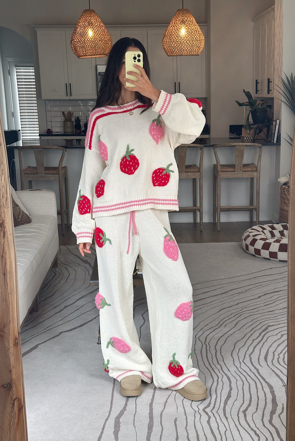 A woman takes a mirror selfie wearing a cozy white knit pajama set featuring a strawberry print and pink striped trim, set in a modern living room with wooden pendant lights and a gray patterned rug.