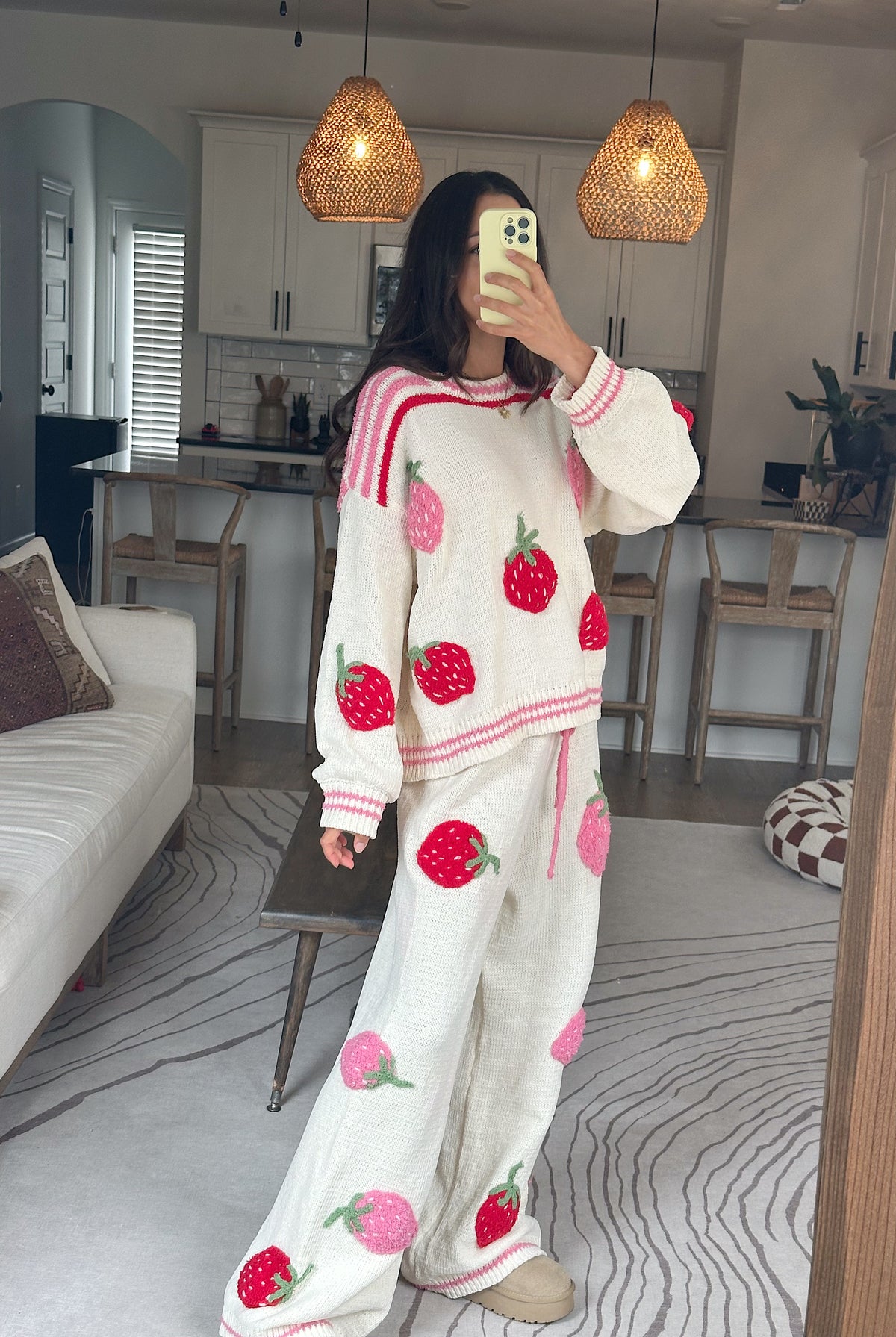 A woman takes a mirror selfie in a cozy living room wearing a matching cream-colored knit lounge set with pink and red strawberry prints. The set includes a long-sleeve sweatshirt and wide-leg pants.