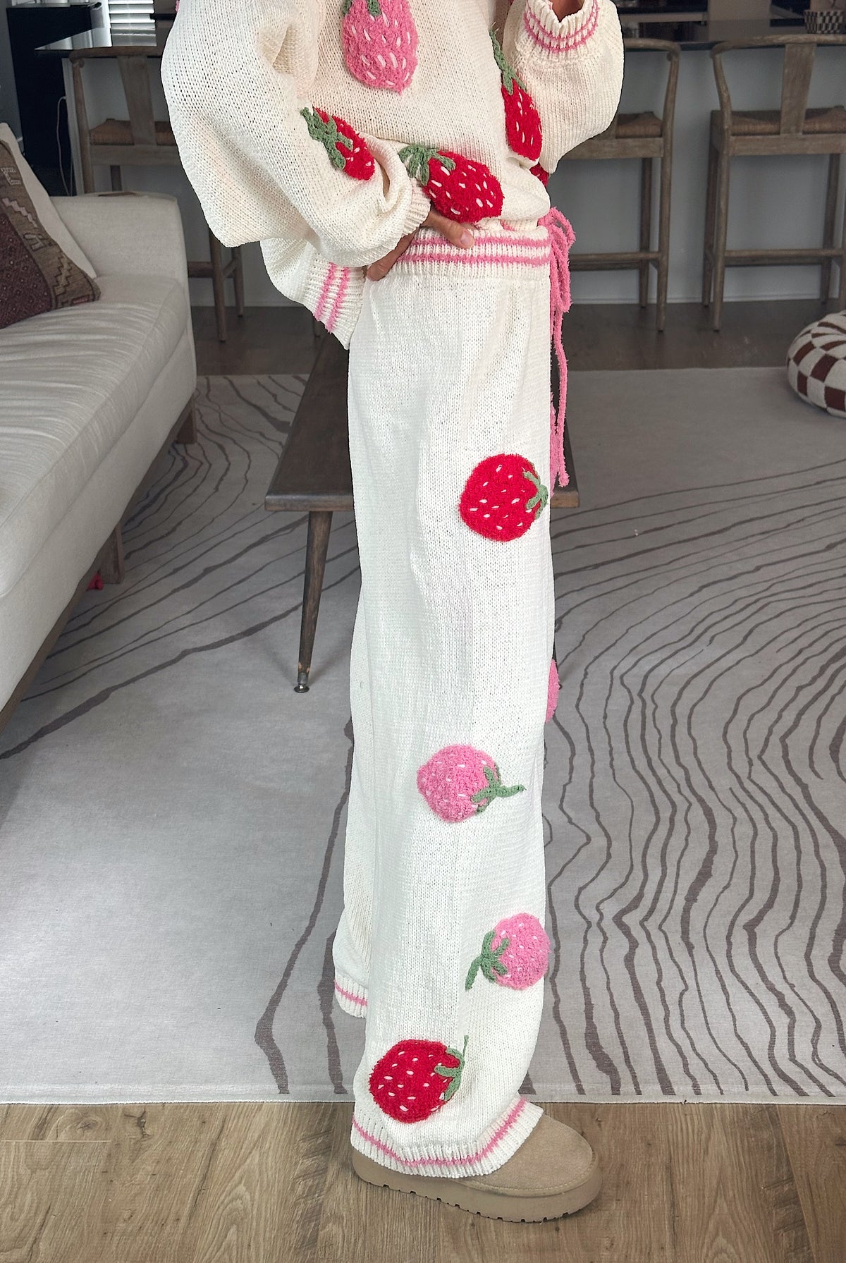 A person wearing a cream knit sweater and matching wide-leg pants with large strawberry appliqués, standing in a living room with a patterned rug and sofa visible.