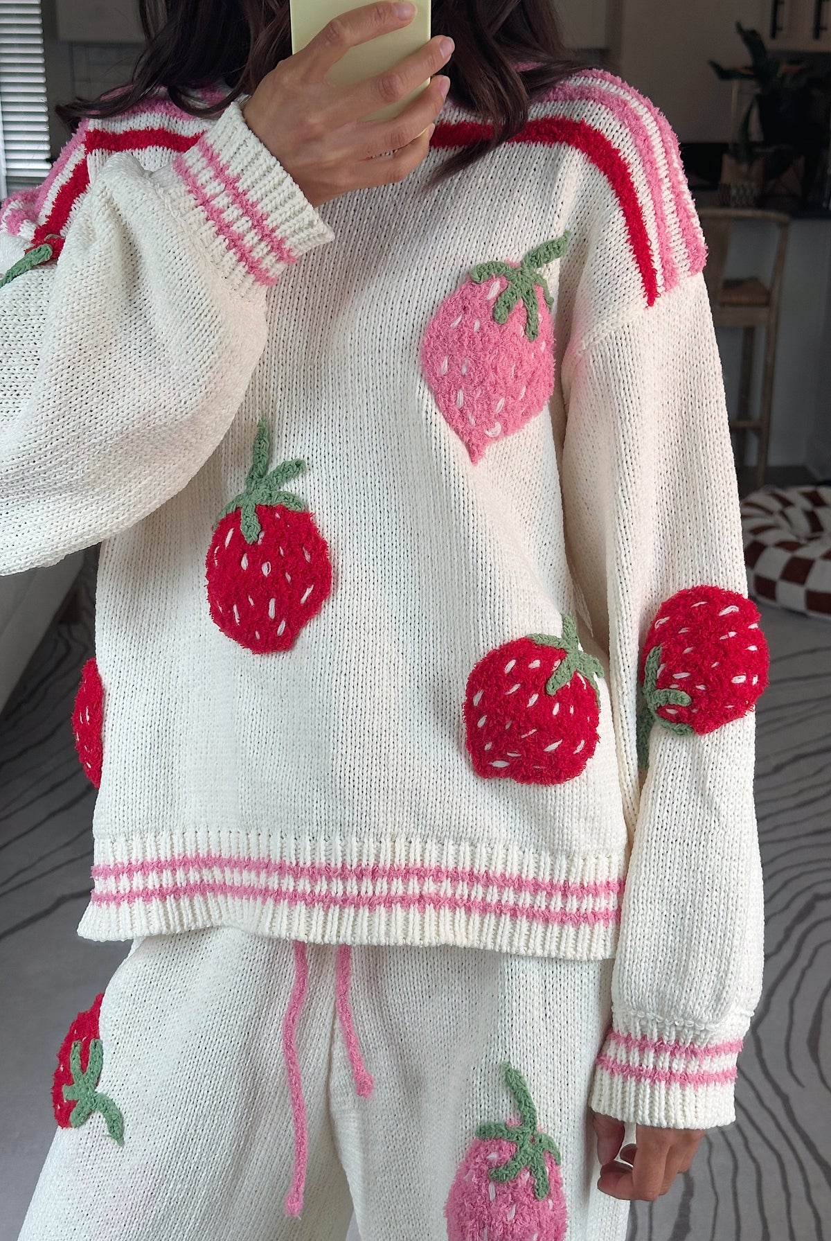 A person wearing a cream knit sweater and matching pants with large red and pink strawberry appliqués, photographed indoors in a home setting.