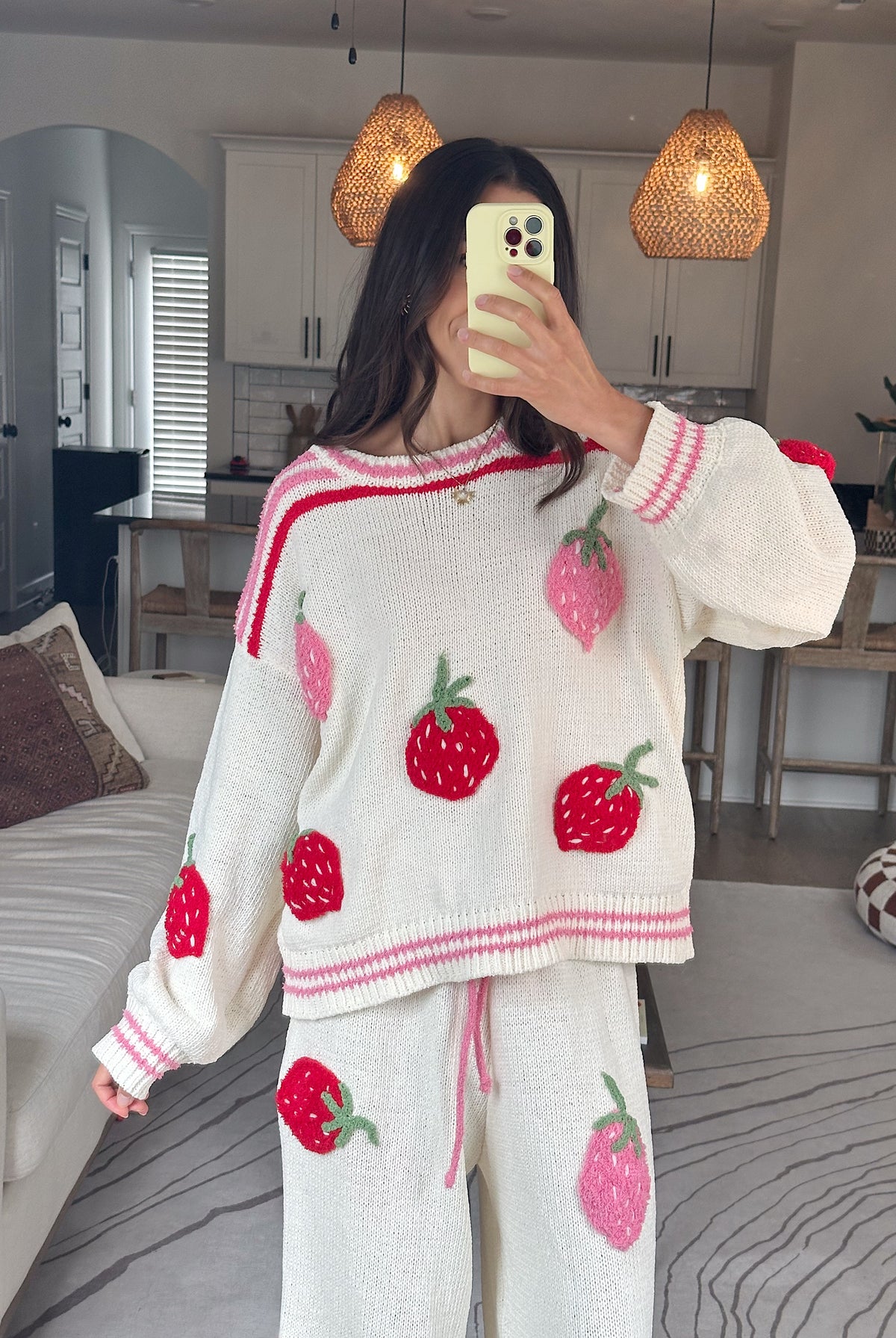 A woman takes a mirror selfie wearing a cozy white knit sweater and matching sweatpants set, both decorated with large red and pink strawberry motifs. The background shows a modern living space with pendant lights, a dining table, and a sofa.