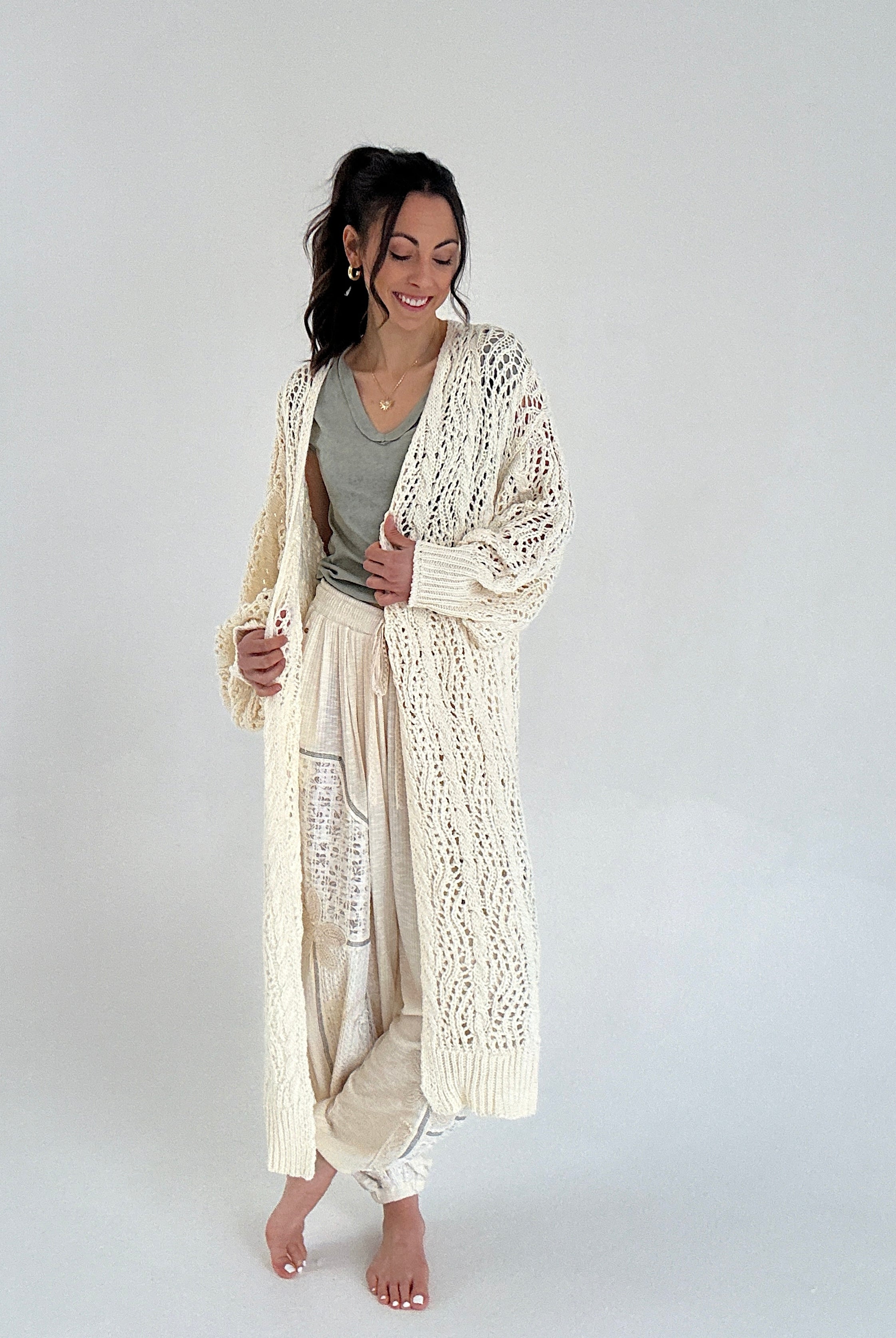 Woman wearing a long, light-colored cardigan over a comfy outfit on a plain background