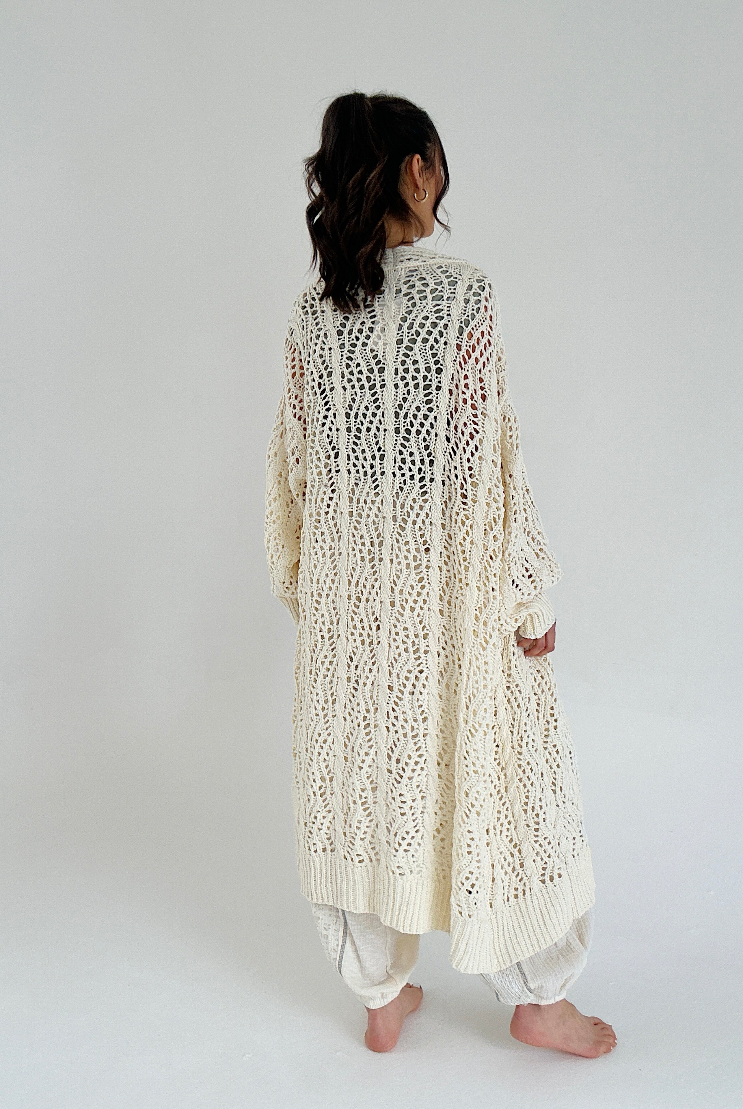 Person wearing a long, cream, knitted cardigan on a plain background showing back