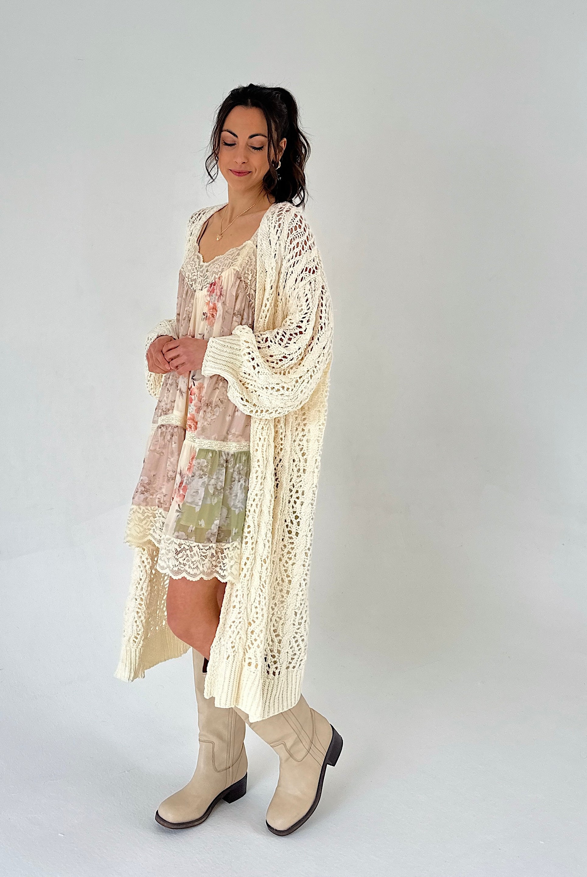 Woman wearing a floral dress and long beige cardigan with white boots on a plain background