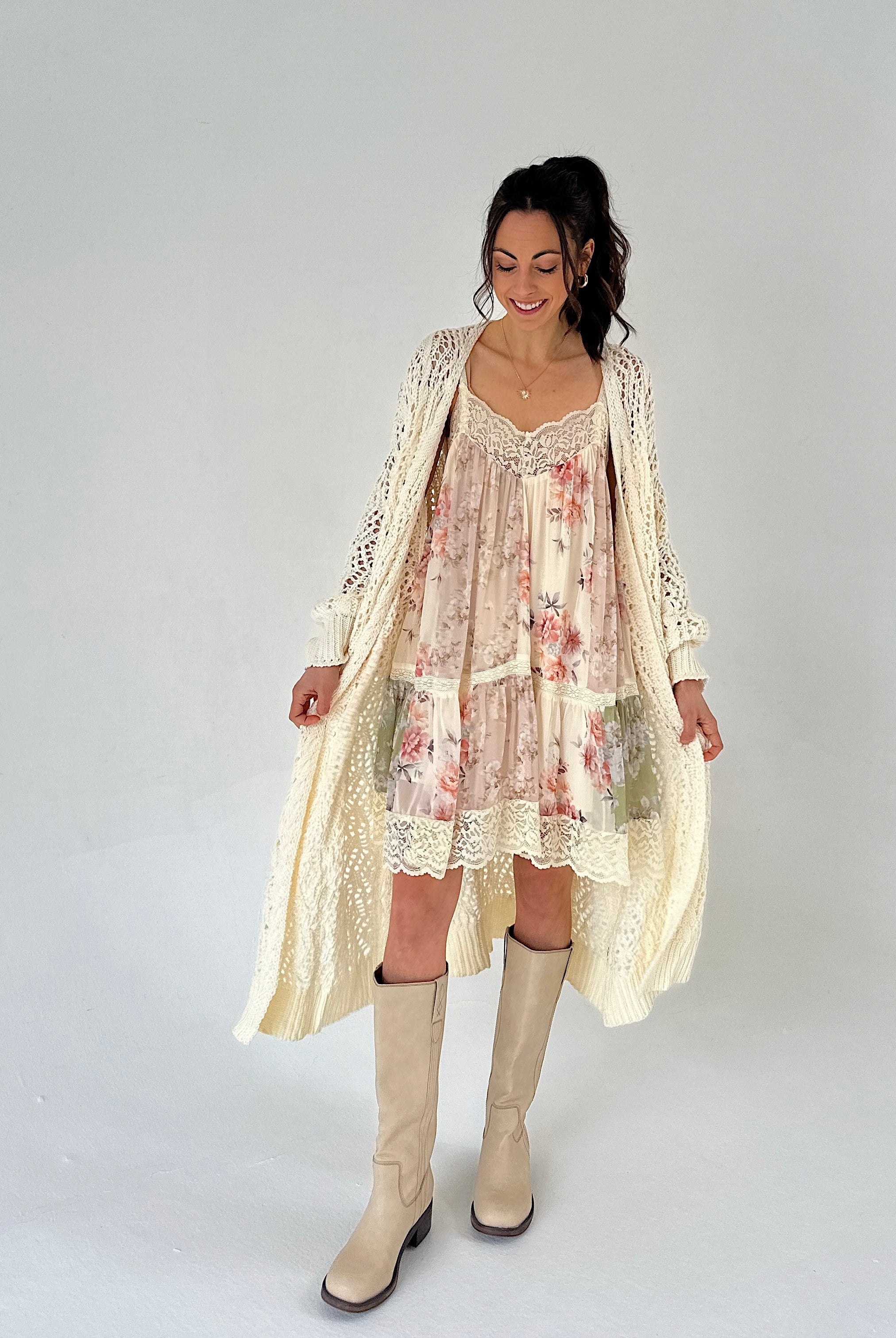 Woman wearing a floral mini dress with a long cream cardigan and knee-high boots on a white background
