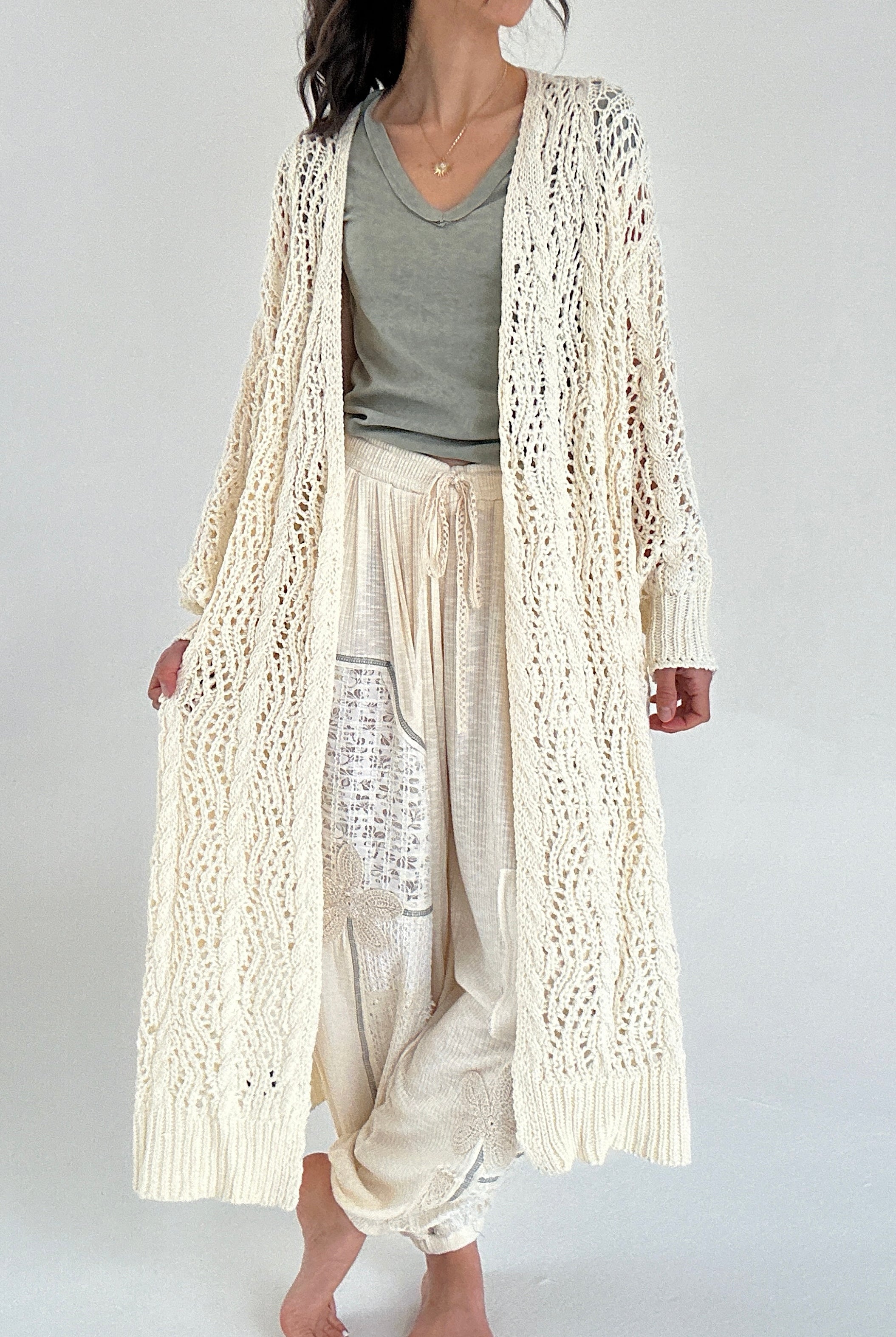 Person wearing a long cream knit cardigan over a green top and light pants on a white background
