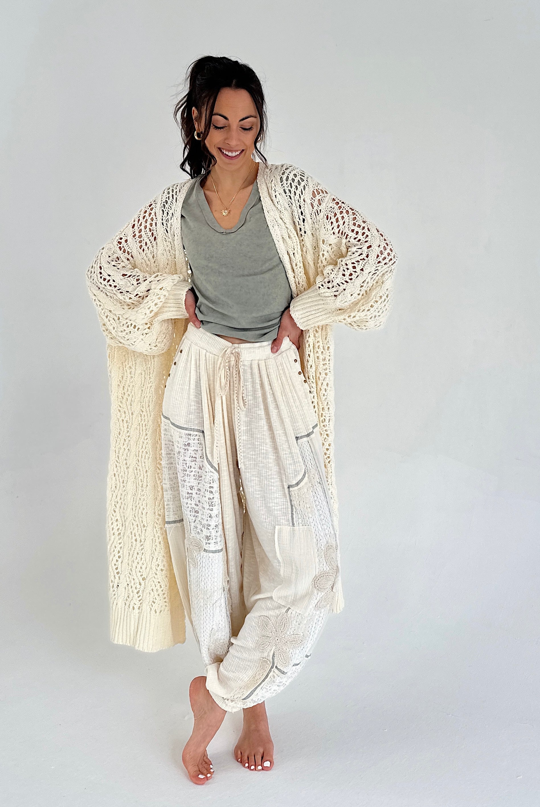 Woman wearing a long, light-colored cardigan over a green/gray top and white pants on a plain background