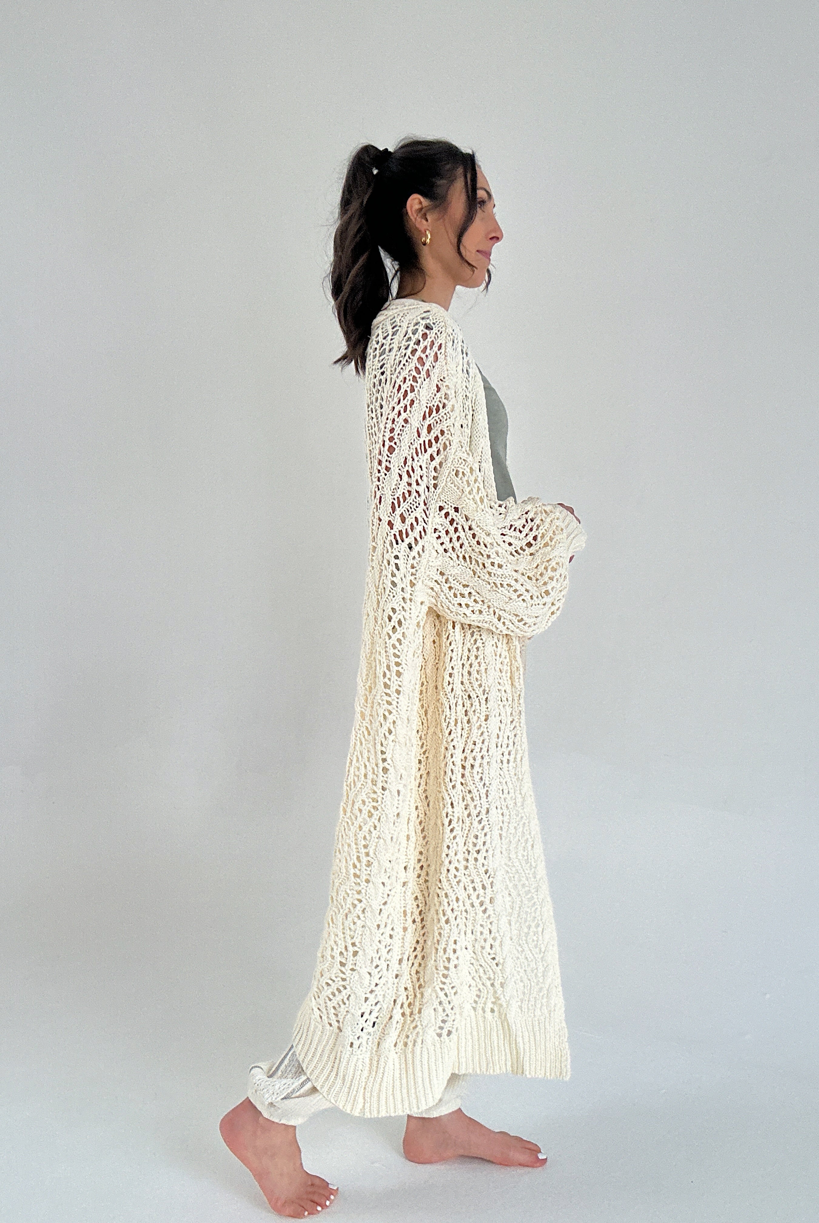 Woman wearing a long, light-colored knitted cardigan against a plain background from side