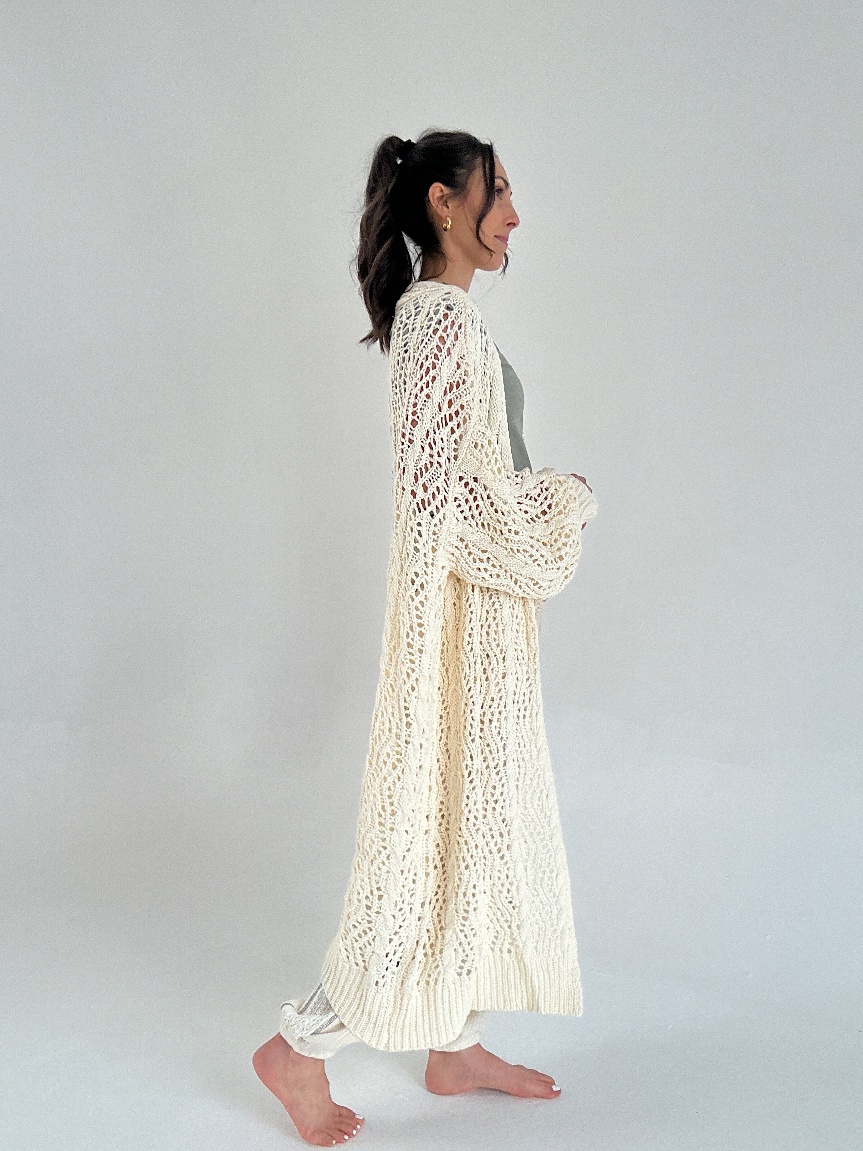 Woman wearing a long, light-colored knitted cardigan against a plain background from side