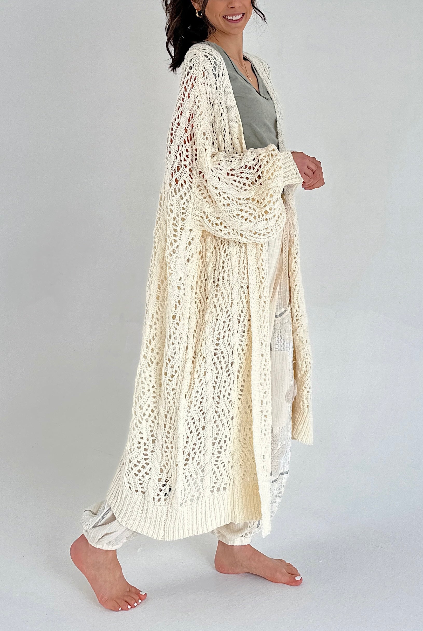 Woman wearing a long, light-colored crochet cardigan on a plain background