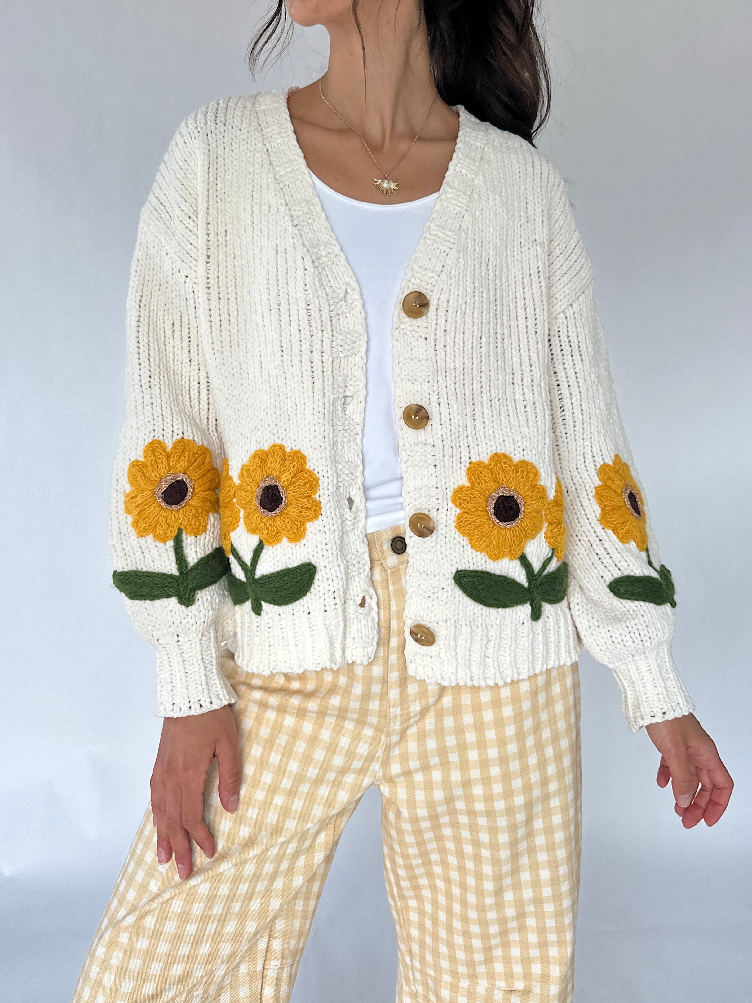 A woman wearing a cream-colored ribbed knit cardigan with large yellow and green sunflower appliqués on the front, paired with a white top and yellow-and-white gingham pants.