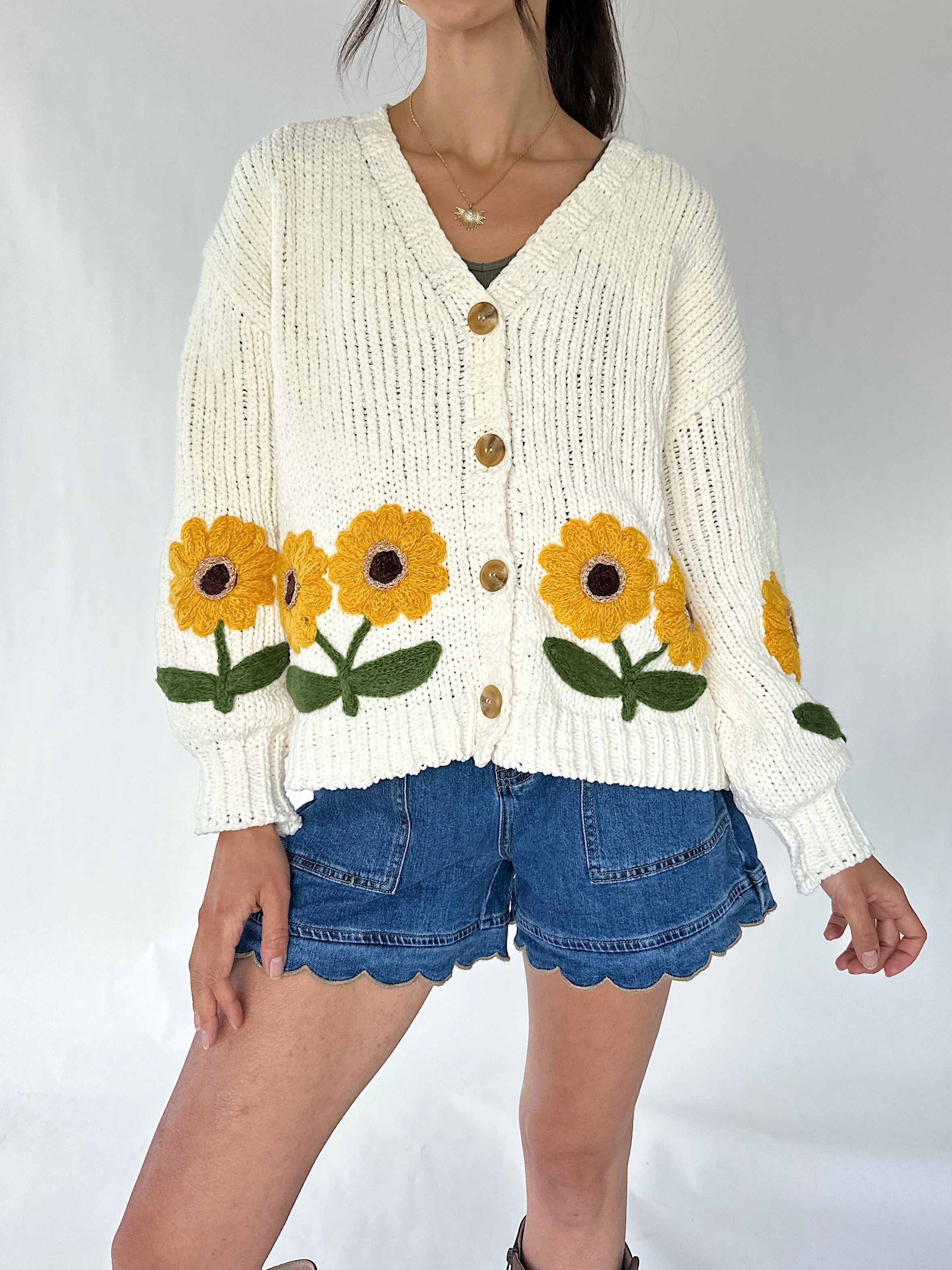 A woman wearing a cream ribbed knit cardigan with yellow sunflower patches and blue denim shorts, photographed against a plain light background.